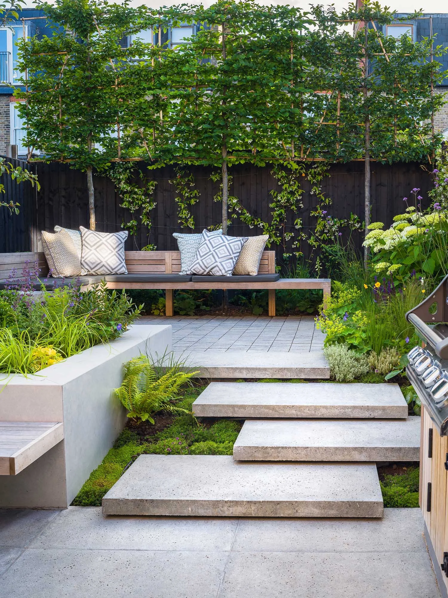 Modern patio with built-in wooden bench, stone steps, and lush planting, with espaliered trees against a dark fence