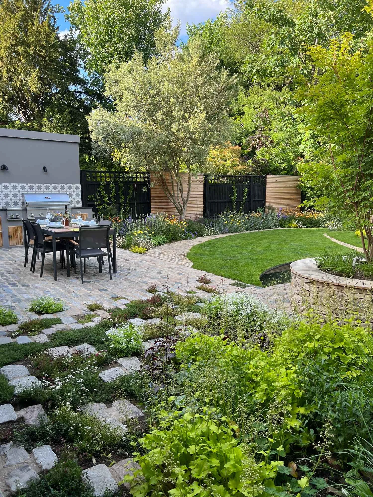 A lush backyard garden with a stone patio, outdoor dining table, and grill, surrounded by trees, shrubs, and flower beds.