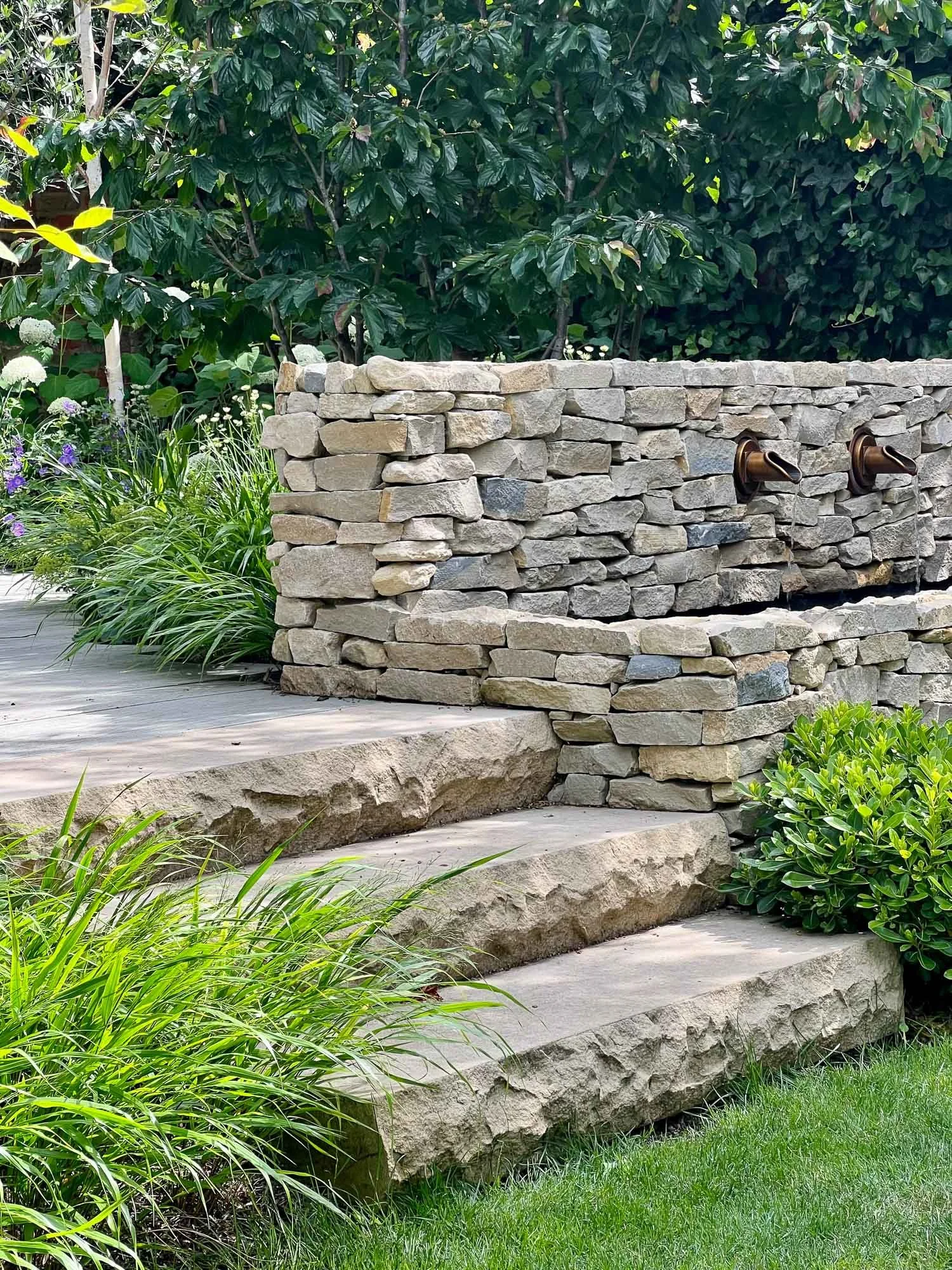 Garden with stone steps, a stone wall with water spouts, and lush green foliage.