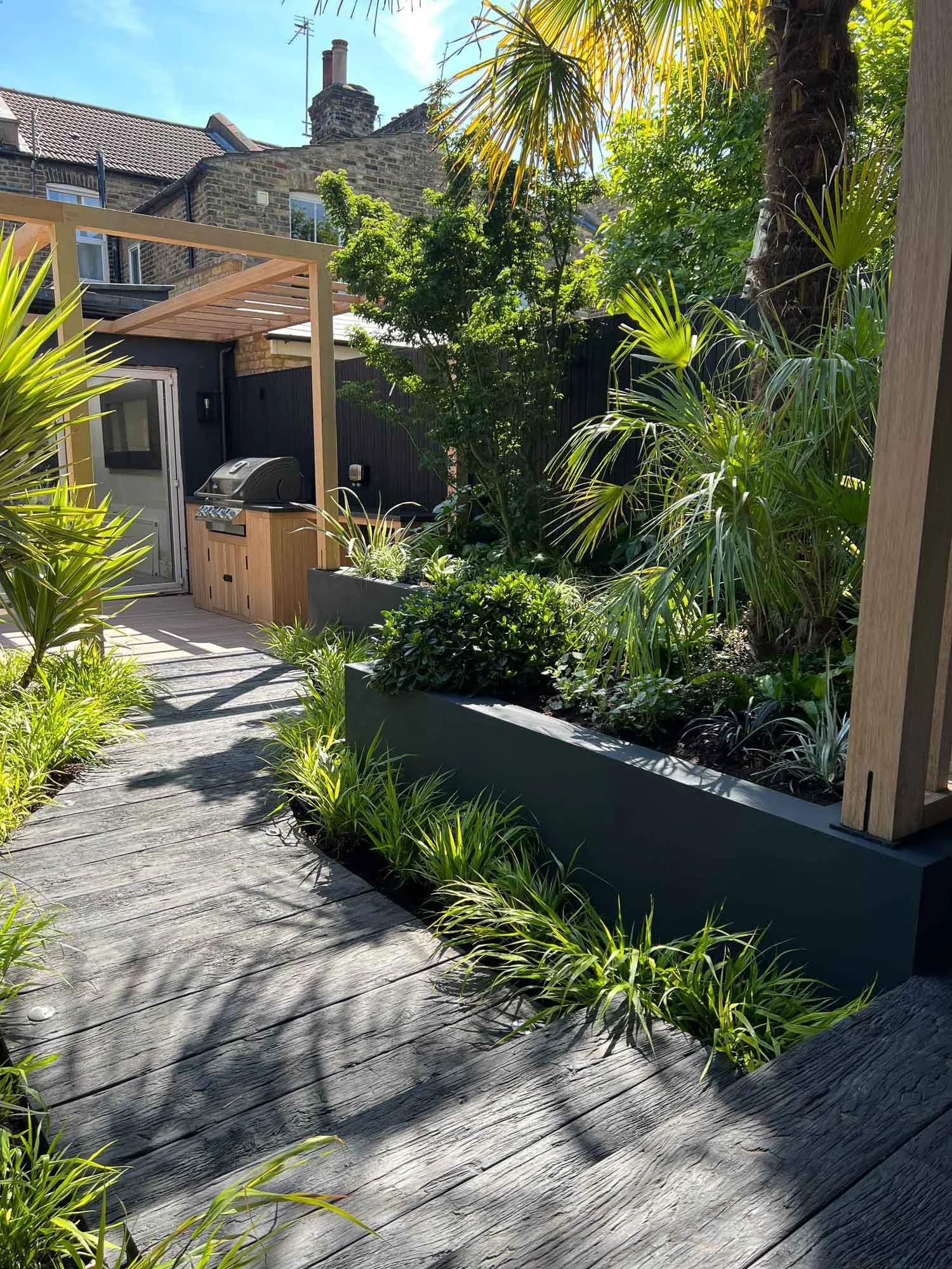 Backyard garden with wooden pathway, tropical plants, palm trees, and a built-in barbecue grill.