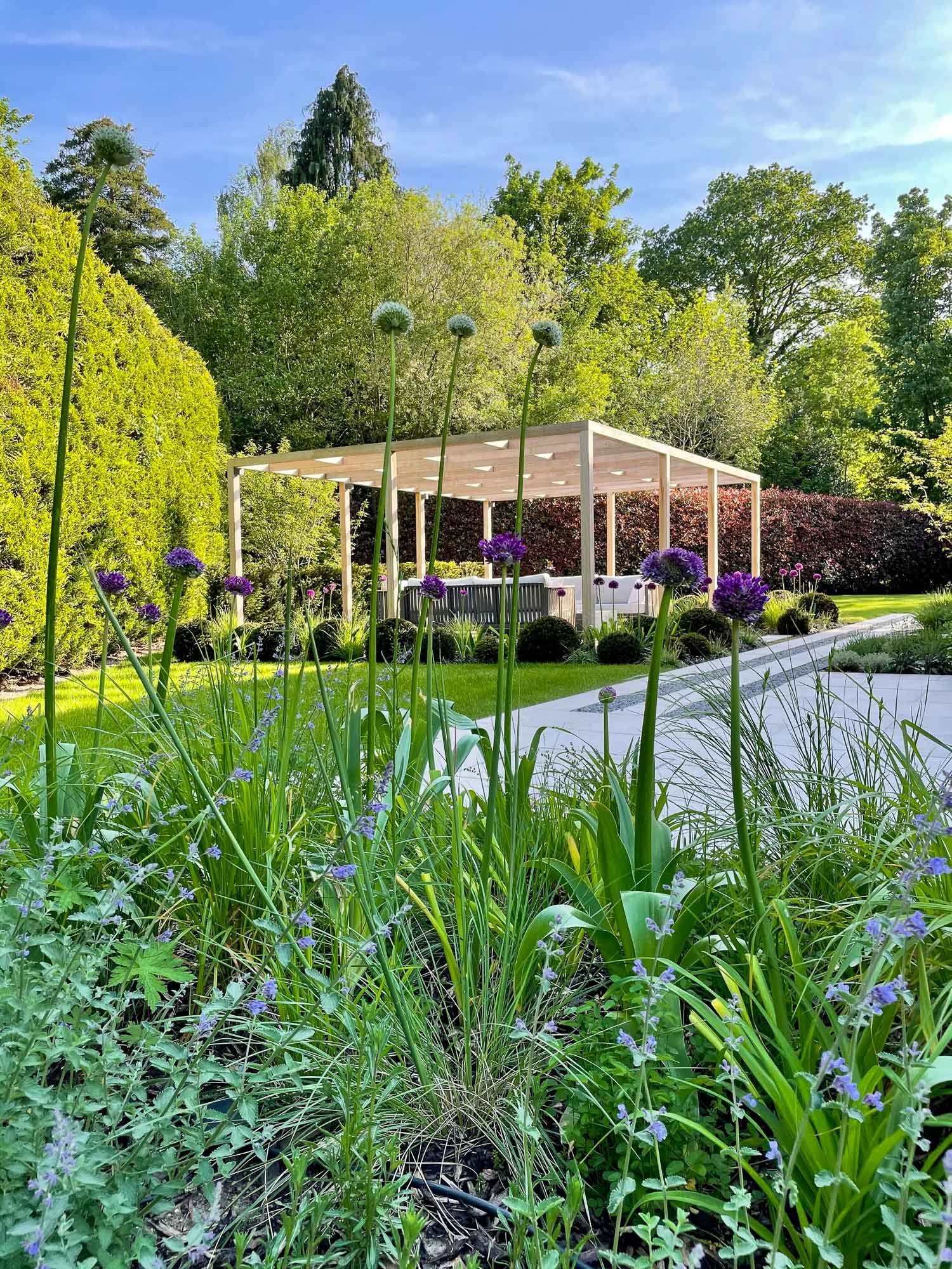 A lush garden with tall purple and white flowers in the foreground, neatly trimmed bushes, a white gazebo in the middle, stone pathway, and green trees under a clear blue sky.