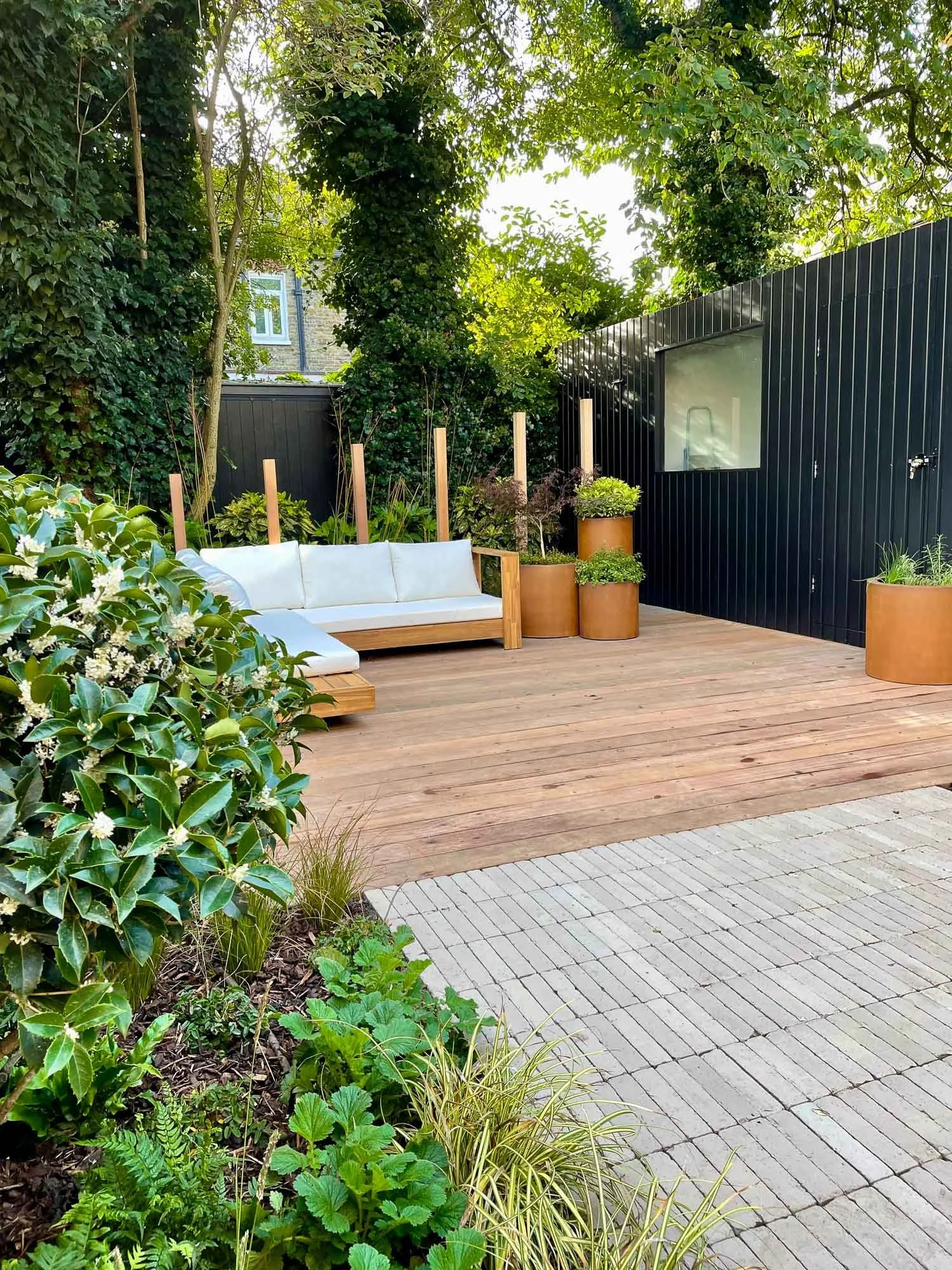A cozy outdoor patio with wooden flooring and potted plants, including a white-cushioned wooden bench, surrounded by lush green trees and bushes, enclosed by black fences, and a window on the black wall.