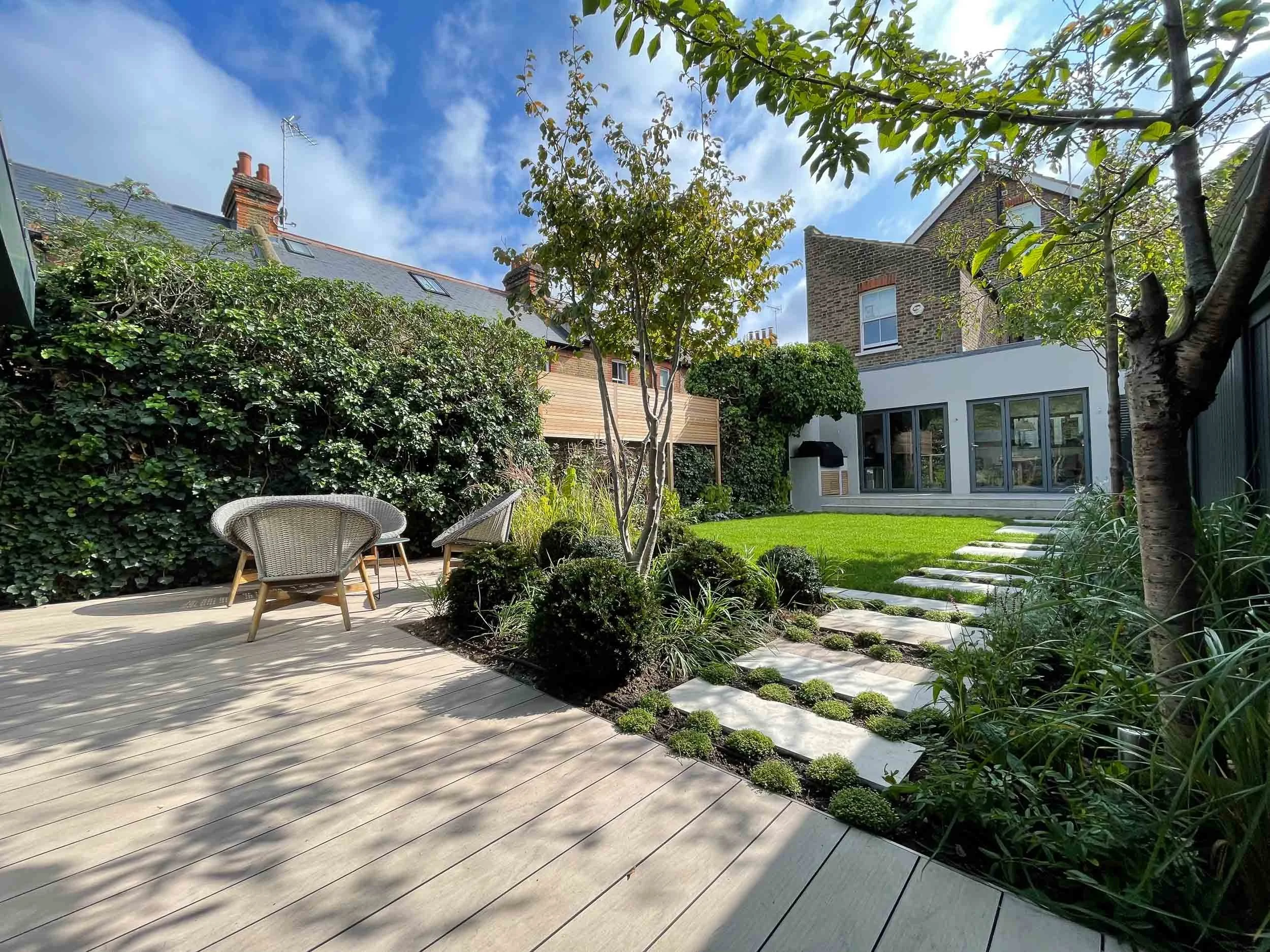 A modern backyard garden with wooden patio, outdoor chairs, lush green grass, trees, and a house with glass doors on a sunny day.