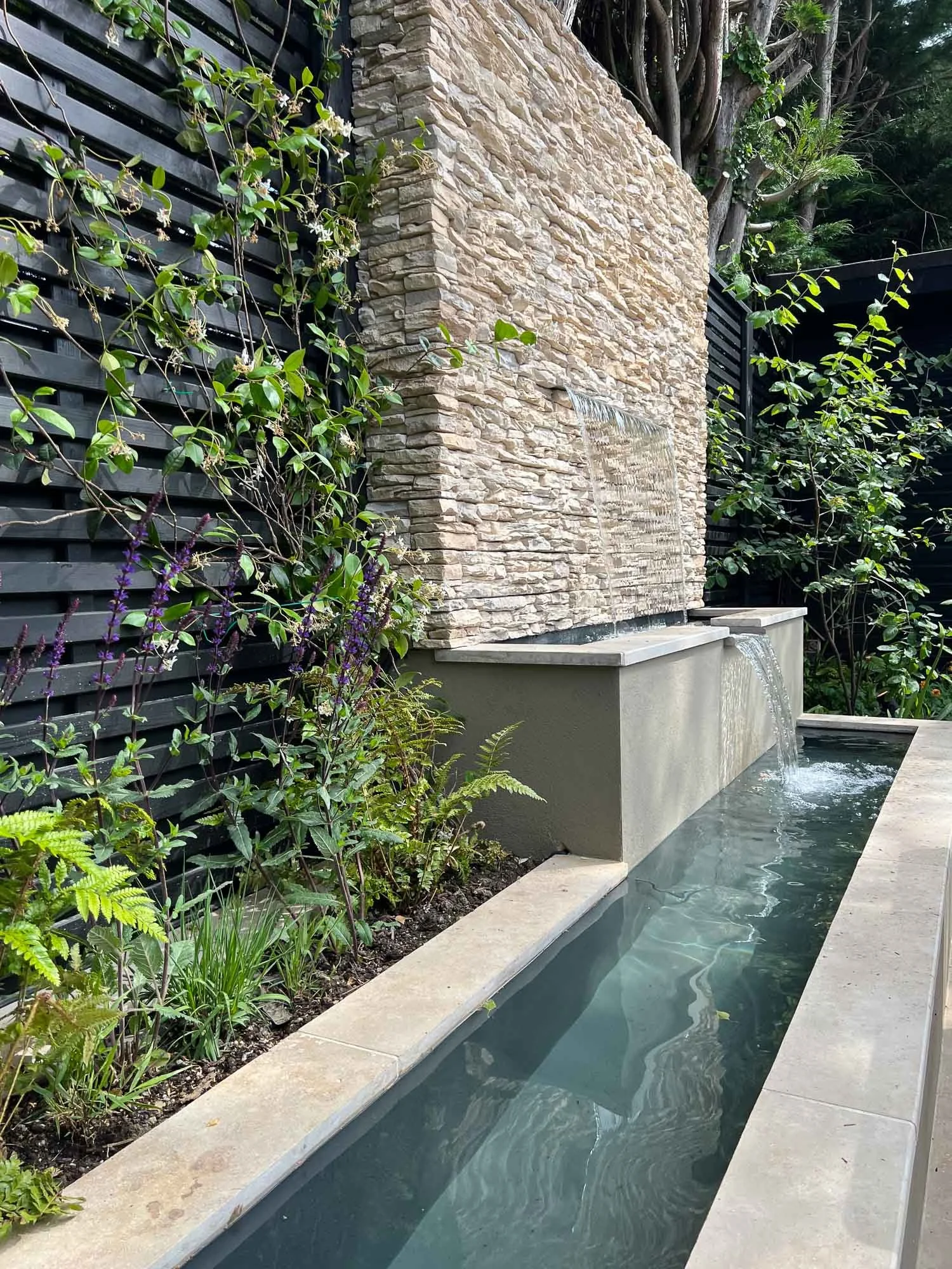 Outdoor water feature with flowing water cascading from a stone wall into a narrow concrete pool, surrounded by greenery and a black slatted fence.