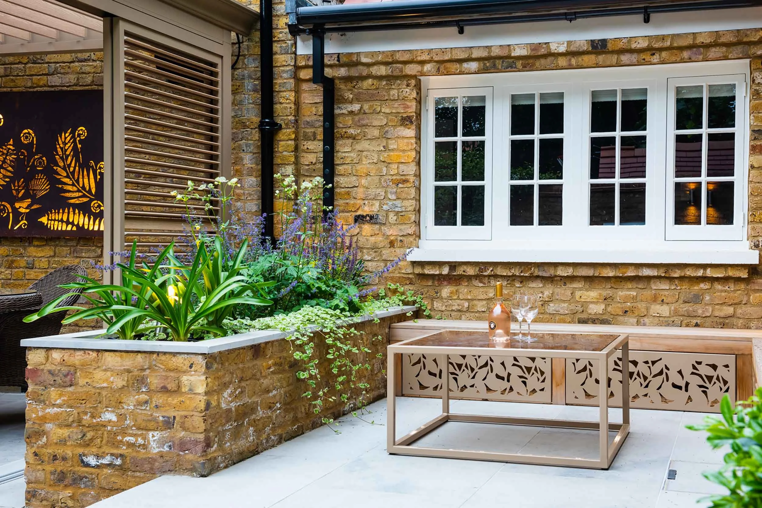 Timber garden bench with laser cut metal panels and a coffee table on a stone patio by a white sash window and planter