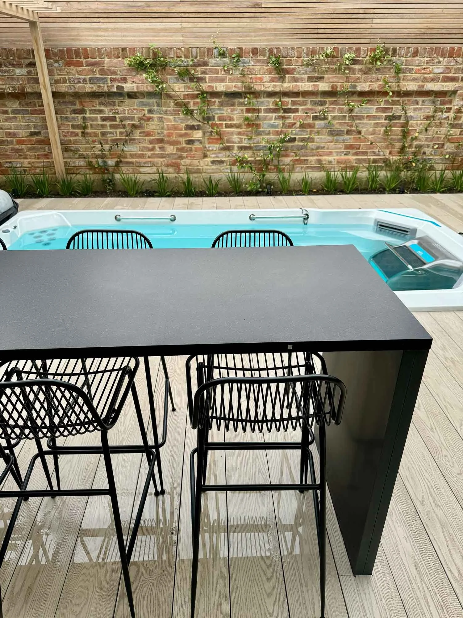 Outdoor patio with a swimming pool, black barstools around a black table, brick wall with green climbing plants in the background.