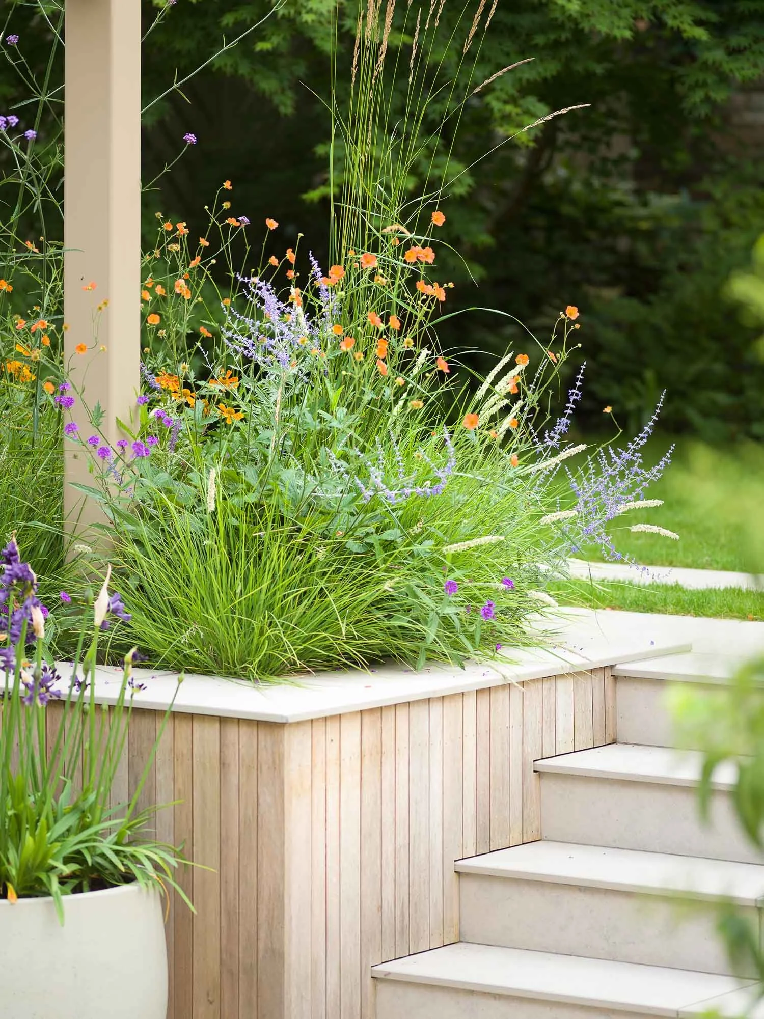 Raised wooden planter box overflowing with tall grasses and orange and purple wildflowers next to stone steps