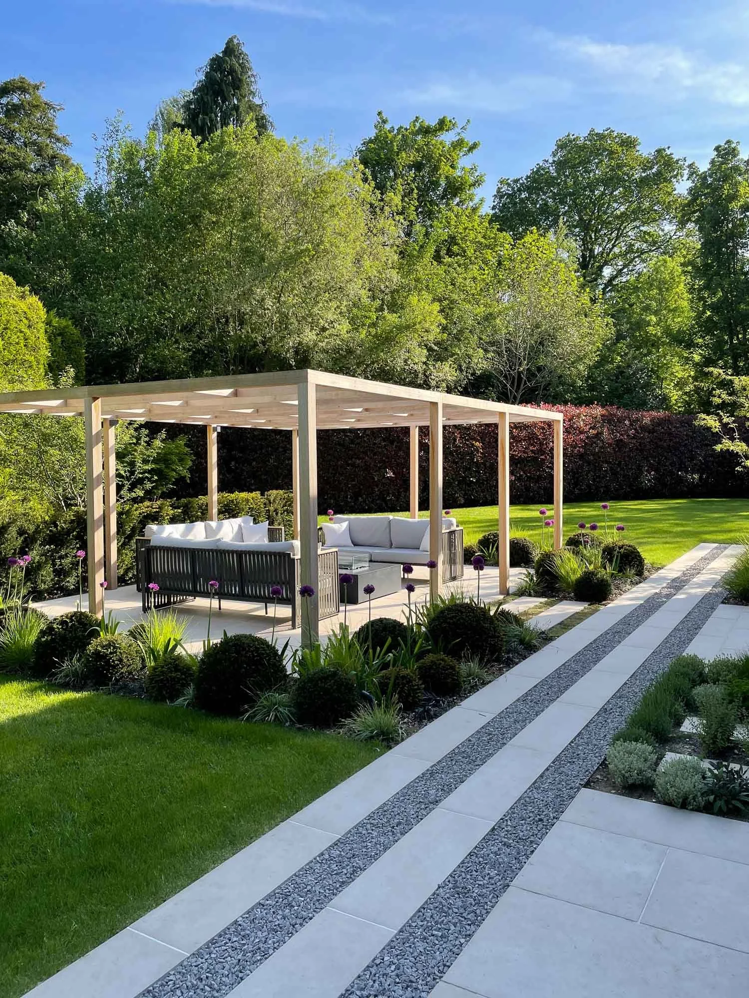 A backyard patio with a wooden pergola, outdoor seating, and a manicured garden with vibrant green trees and purple flowers under a clear blue sky.