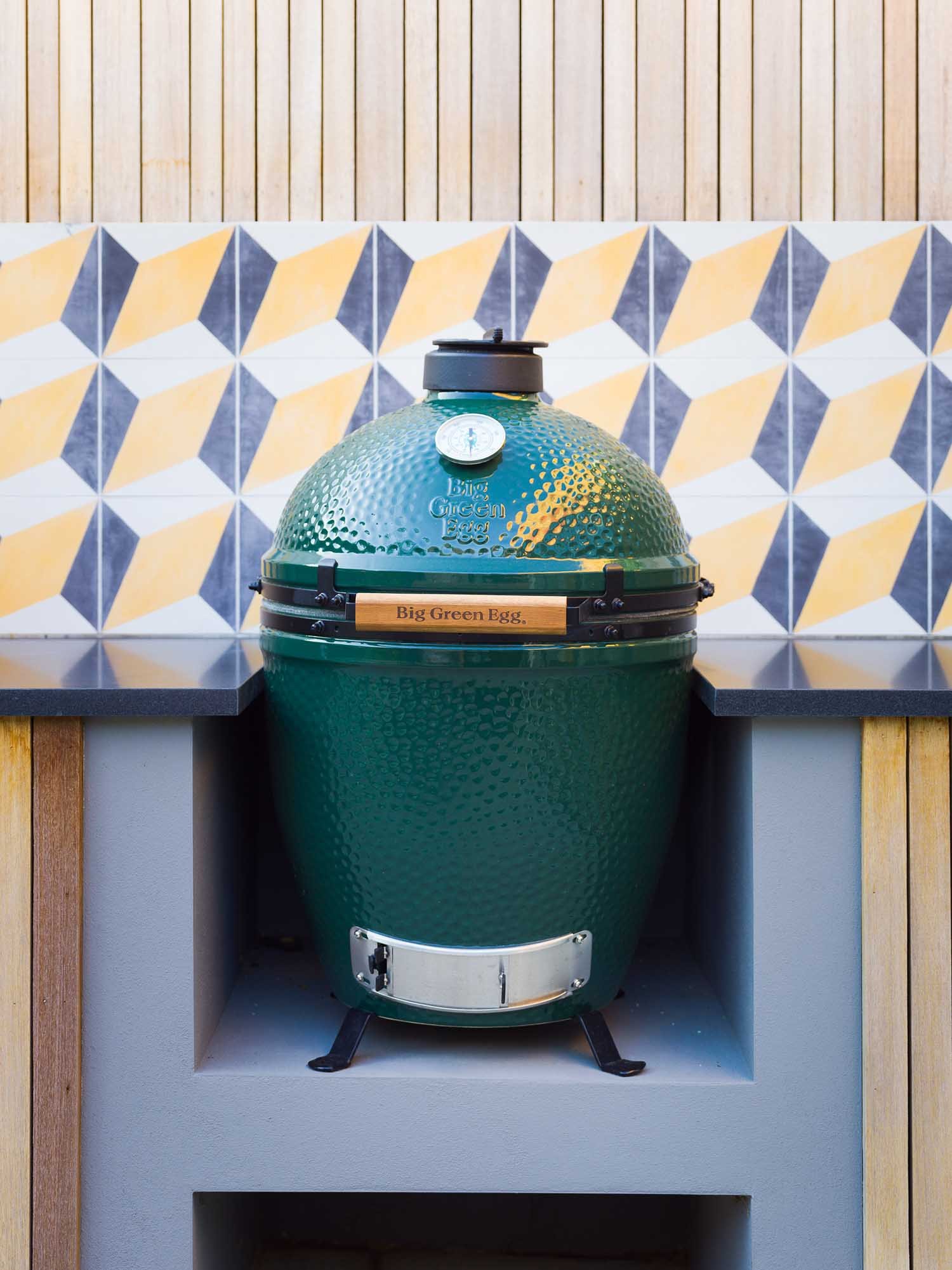 Close-up of a green Big Green Egg grill on a dark counter with yellow/grey geometric tiles and vertical wooden slats