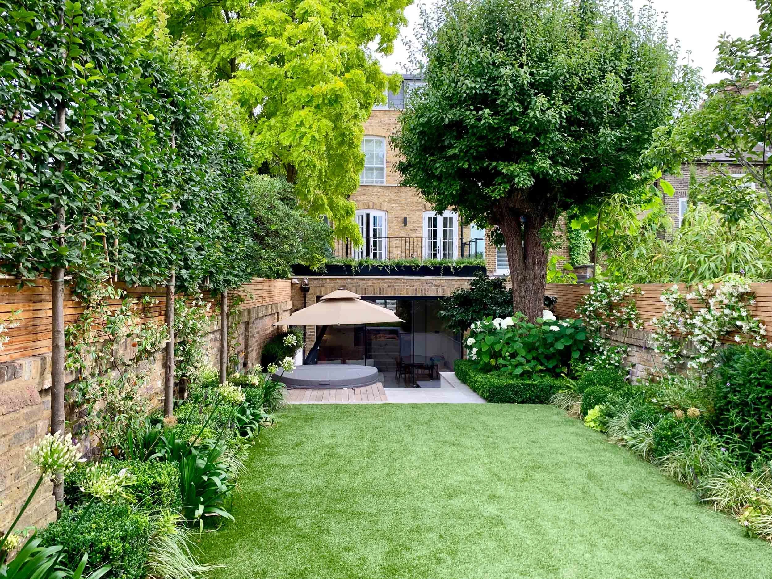 A landscaped backyard with a grassy lawn, tall trees, blooming plants, and a patio area with an umbrella and a hot tub, bordered by brick and wooden fences.