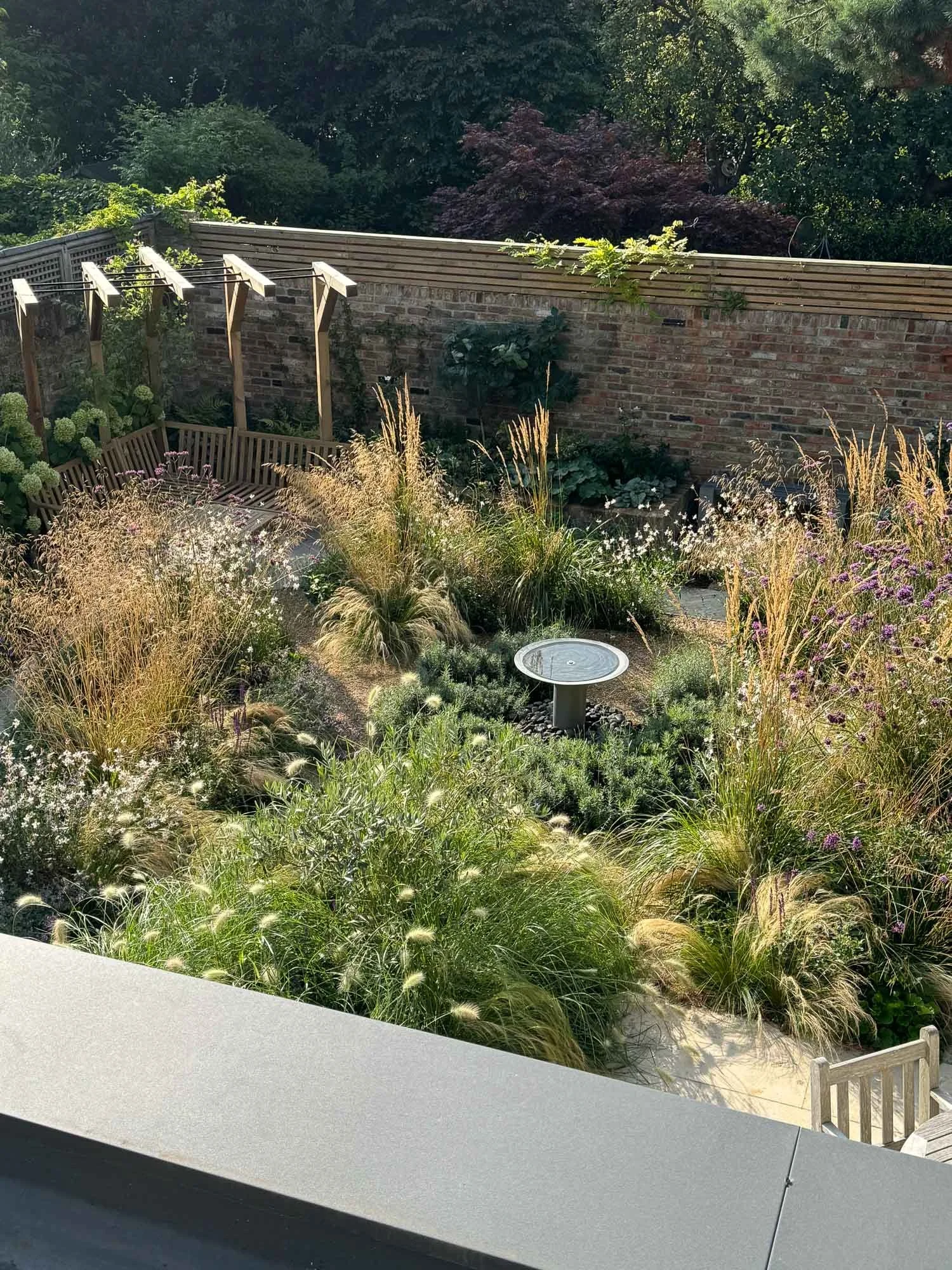A lush garden viewed from above, with a variety of tall grasses, flowering plants, and a small circular birdbath, enclosed by a brick wall and a wooden fence.