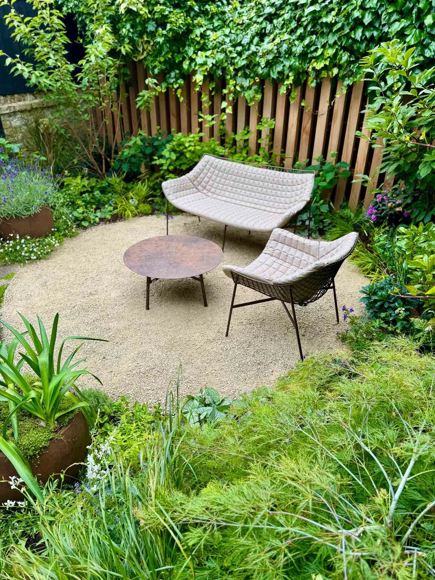A garden patio on light gravel with two quilted chairs and a rusty metal table, surrounded by ferns, grasses, and a vertical wood fence