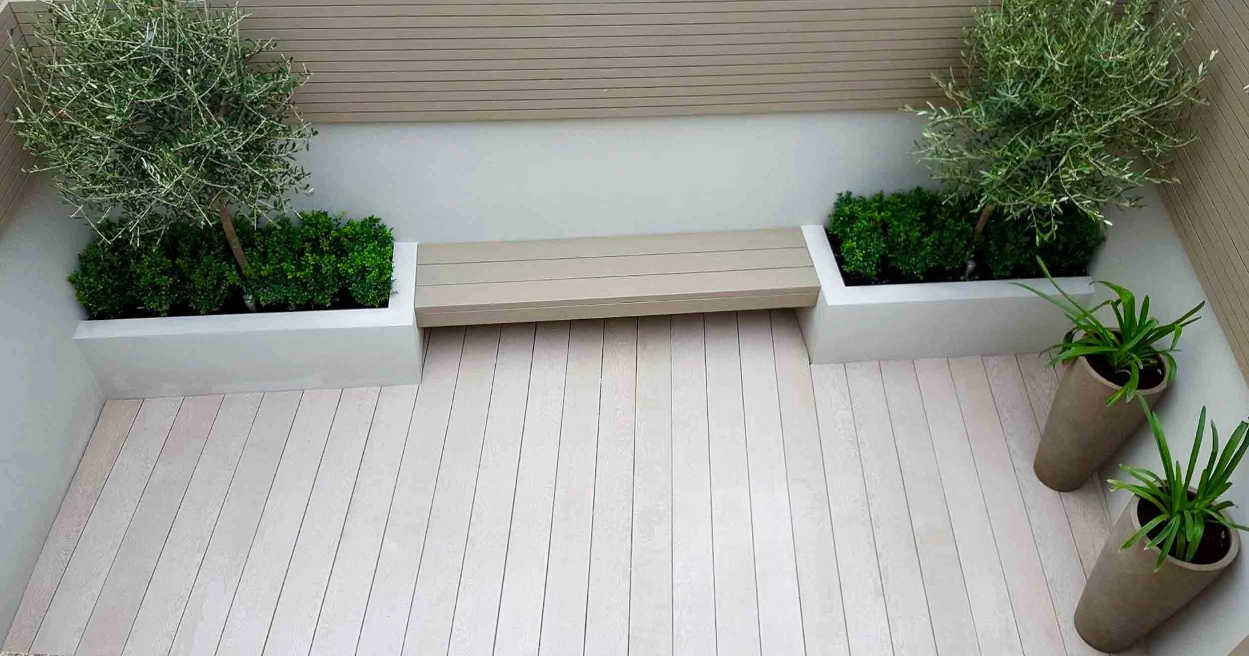 Corner of a balcony with two planters containing green leafy plants and a small wooden bench.