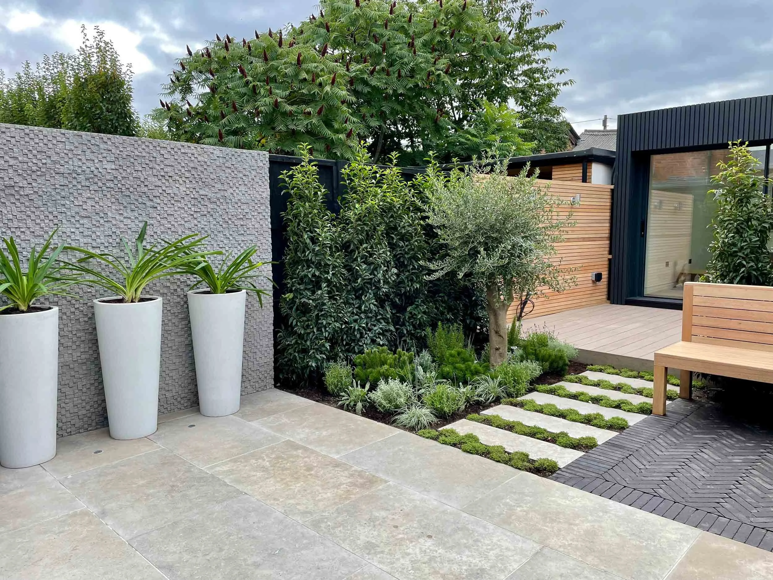 Modern backyard garden with potted plants, shrubs, trees, wooden deck, and a black sliding glass door, under cloudy sky.