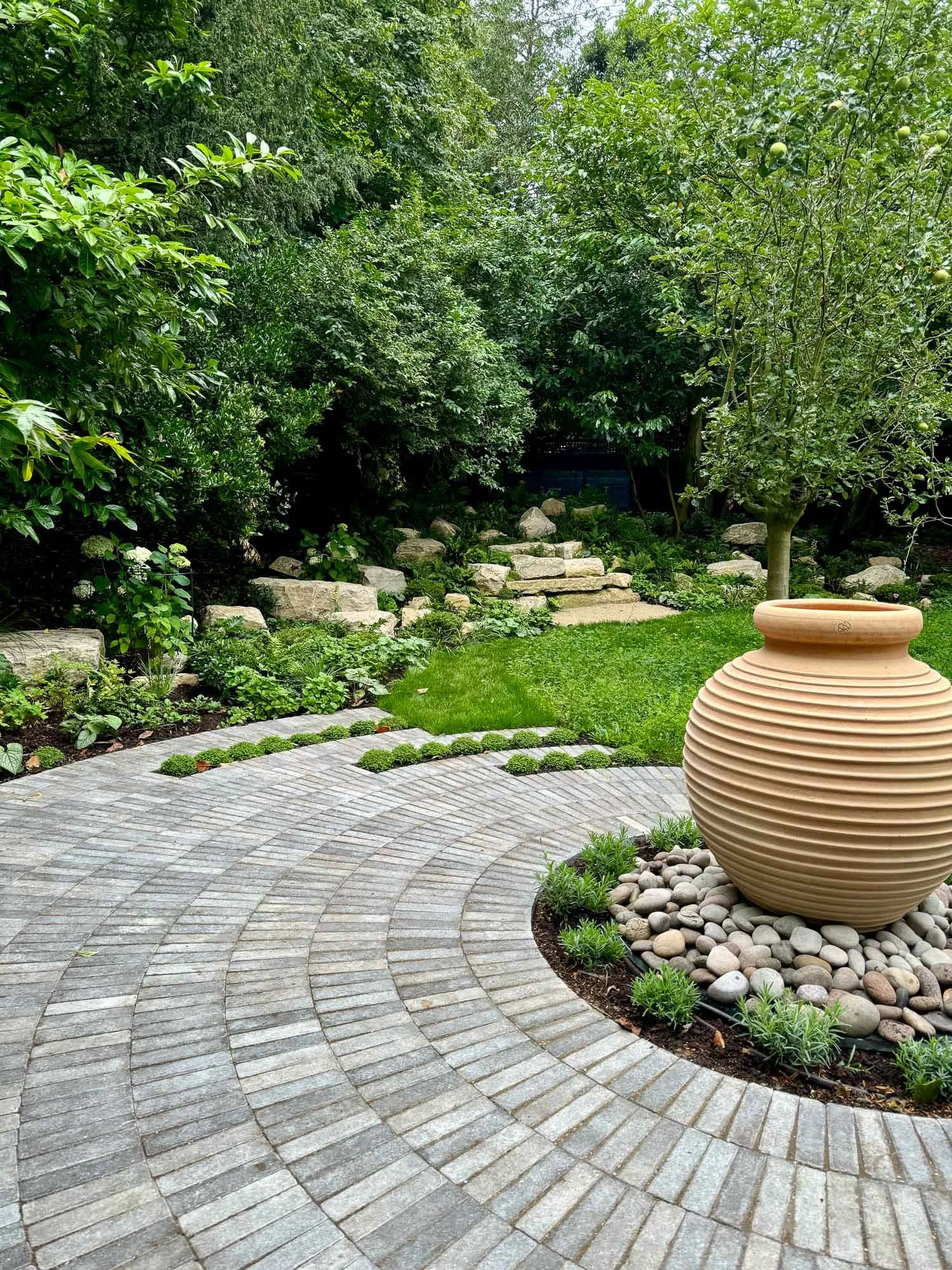 A landscaped garden with a circular brick patio, a large beige ceramic vase surrounded by rocks, and lush green trees and plants in the background.