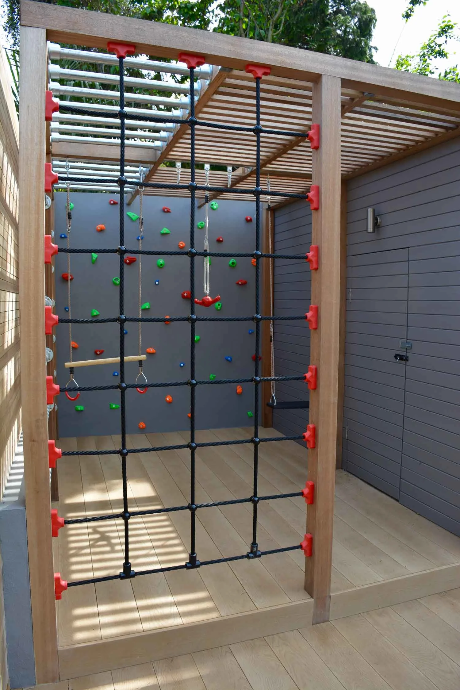 A backyard play area with that includes a climbing wall, a rope net for climbing, and a swing, all set within a wooden framing structure.