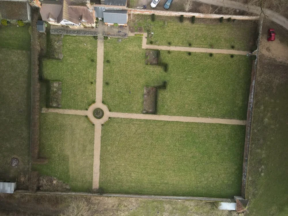 Outdoor patio with wooden pergolas, stone planters, a central tree surrounded by purple flowers, lounge seating, a fire pit, and surrounding greenery under a partly cloudy sky.