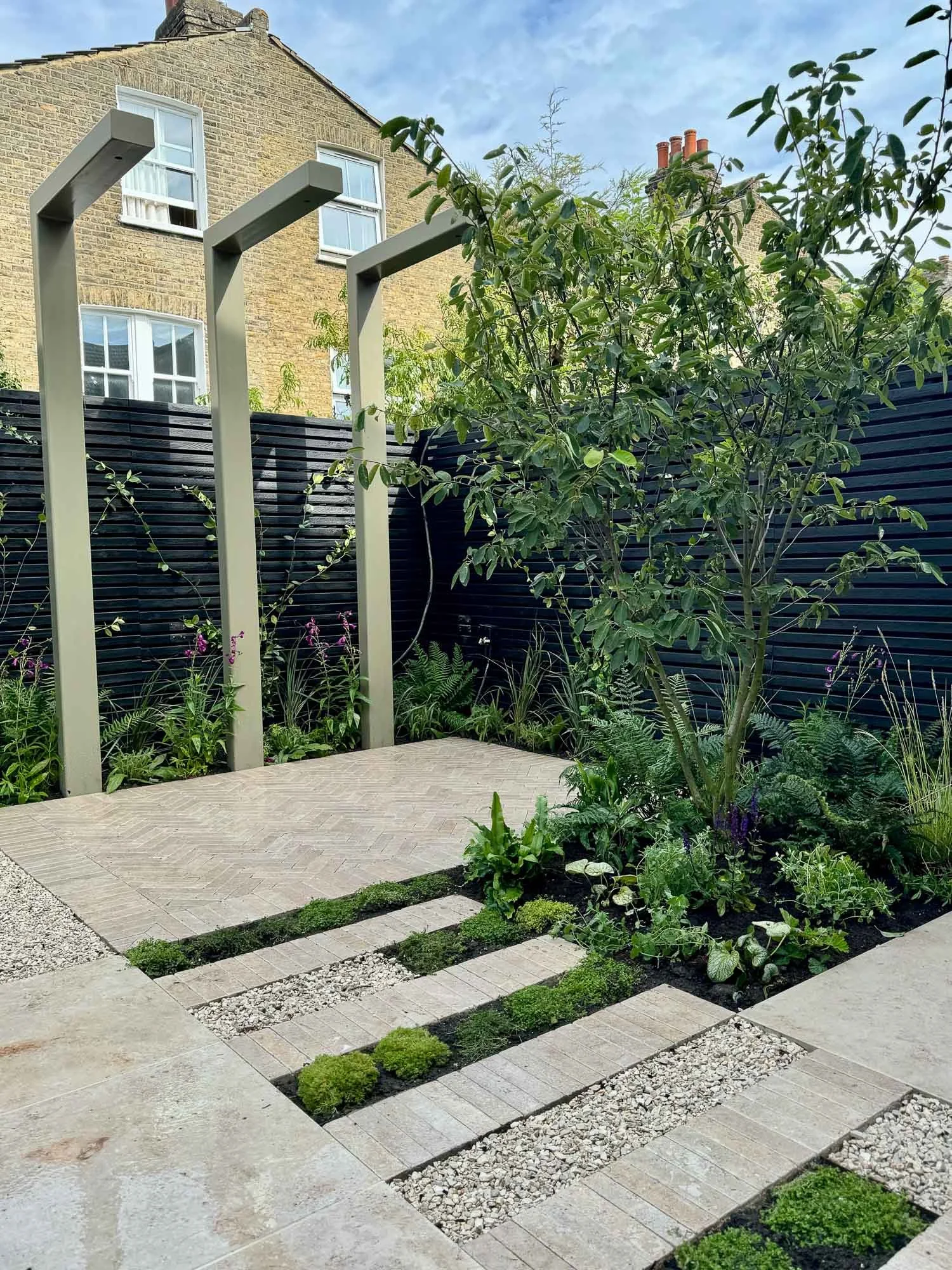 Modern backyard garden with paved patio, small plants, lush greenery, black fence, and three tall, rectangular wooden structures.