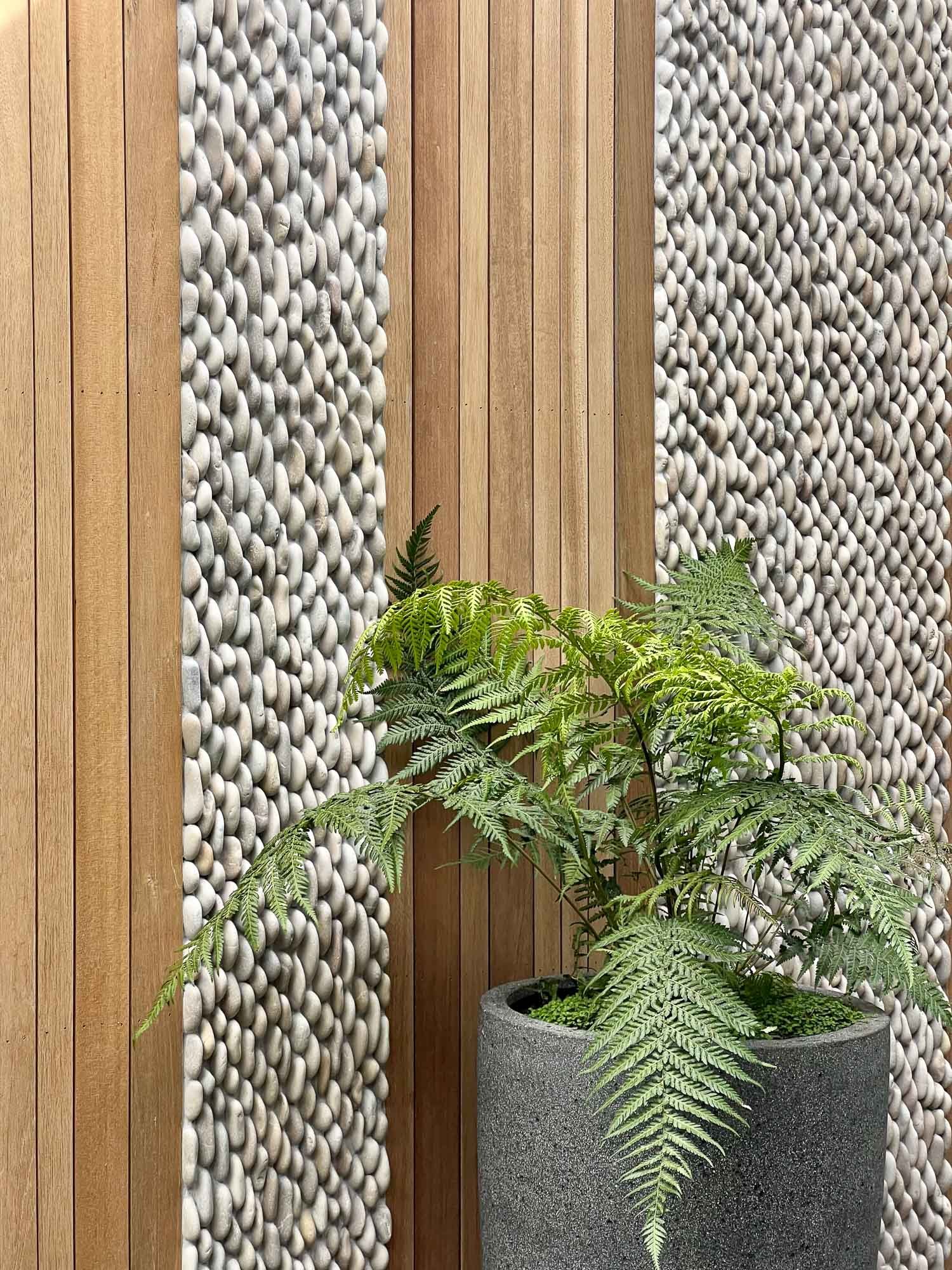 A potted fern plant with green fronds in a gray concrete planter against a decorative wall with vertical wooden slats and stone pebble accents.