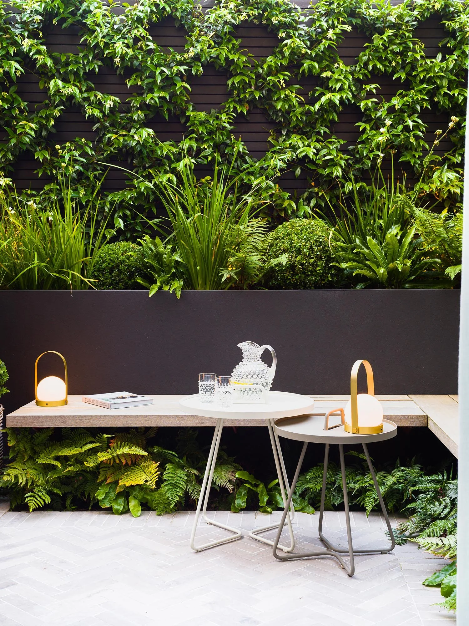 Contemporary courtyard seating area with a light wood bench and white tables set against a wall of climbing plants