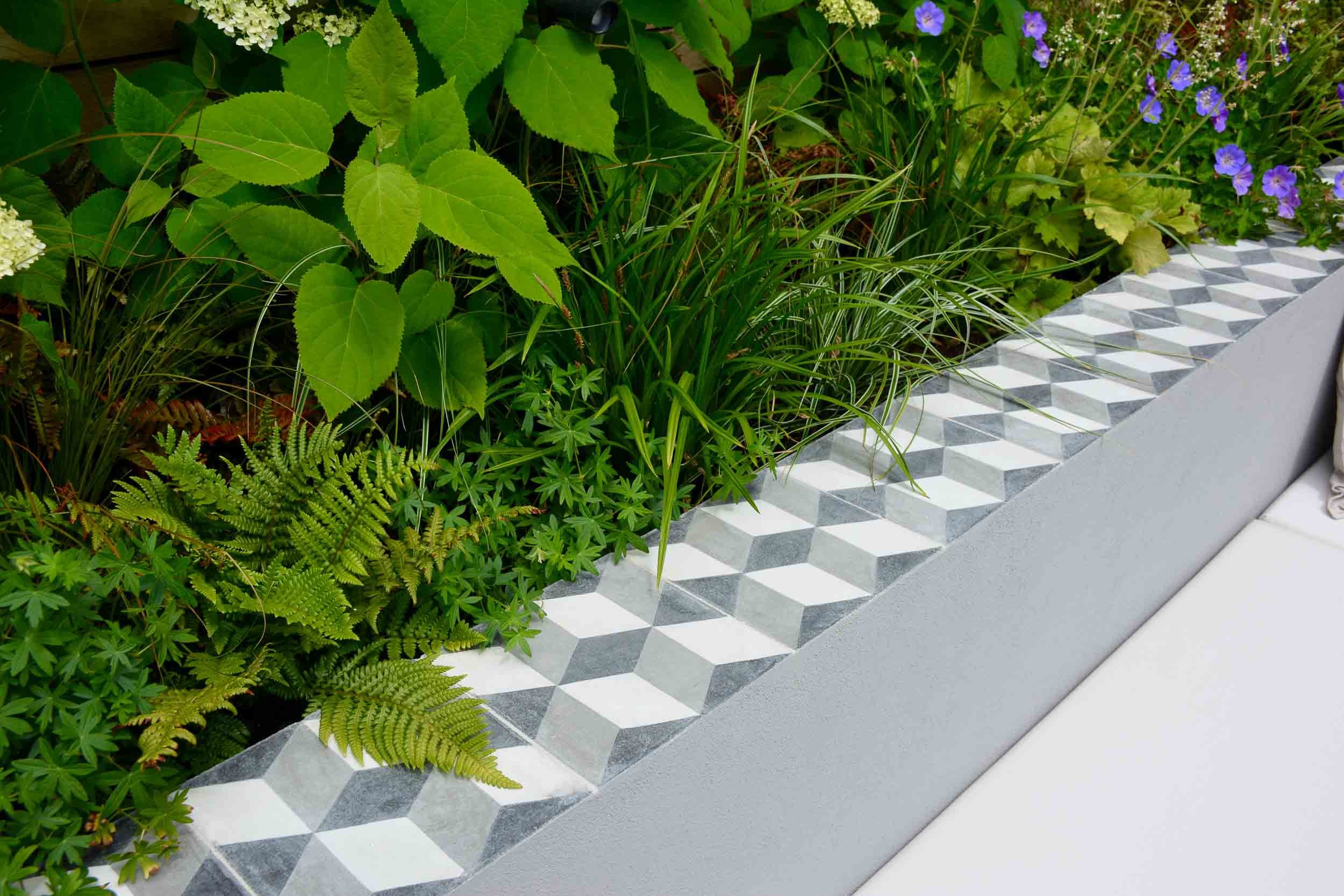 Close-up of a modern concrete garden bed with geometric patterned tiles, lush green plants, and small purple flowers