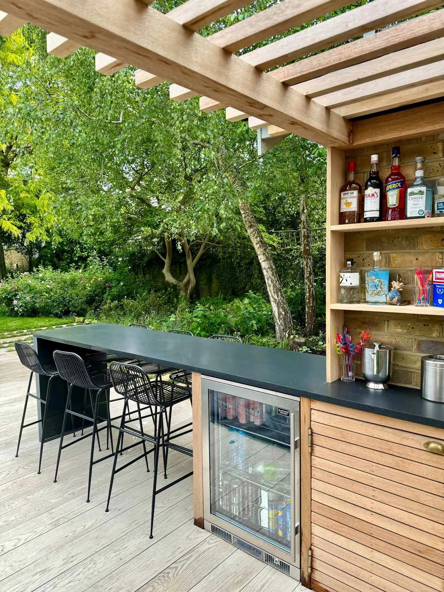 Outdoor bar area with black countertop, bar stools, open cabinet with liquor bottles, a small fridge, and a lush garden with trees and bushes in the background.