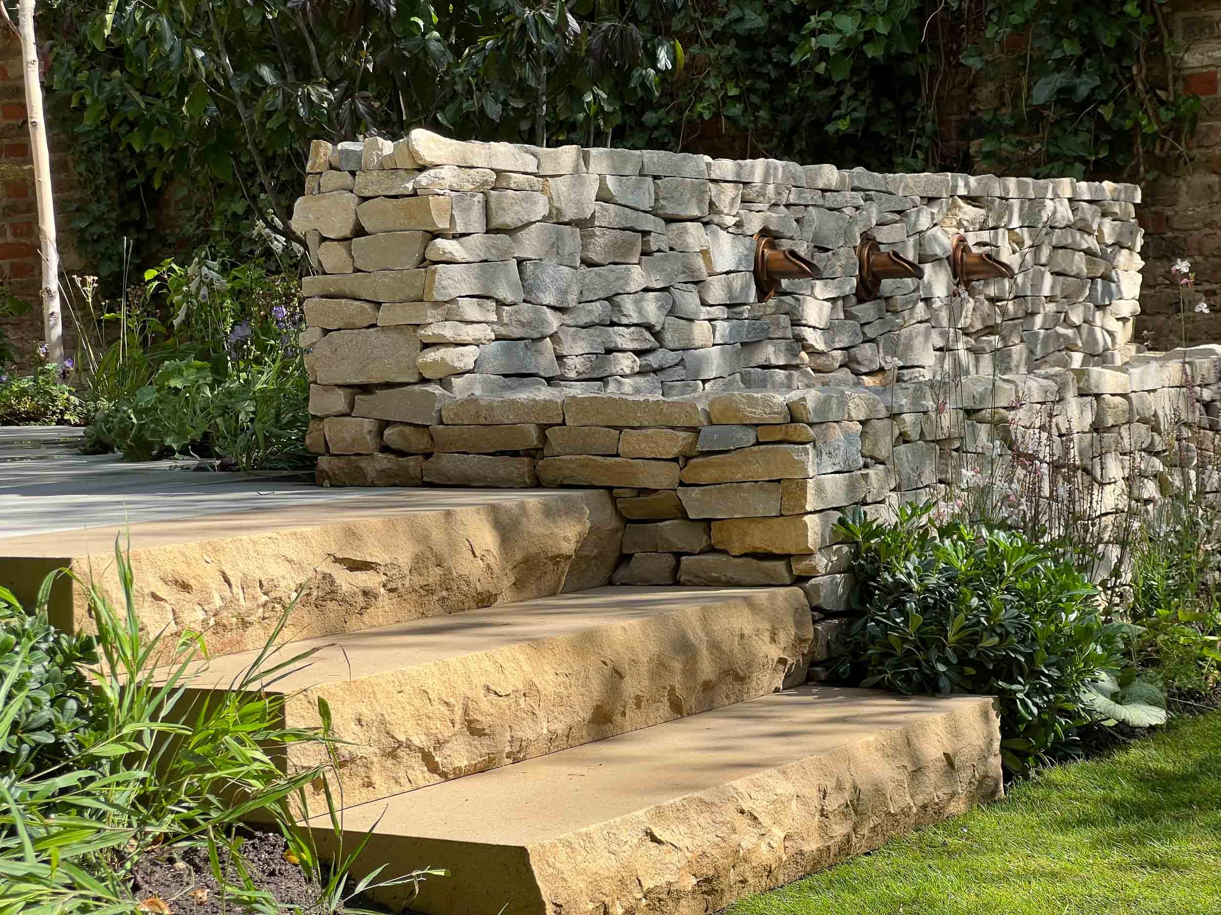 Stone water fountain with three brown spouts in a garden setting, surrounded by greenery and plants.