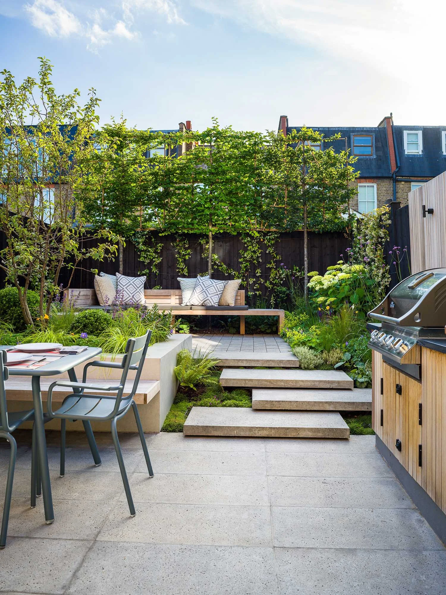 Sunny view of the garden patio, showing the paved dining space, stone steps, wooden bench, and tiered planting