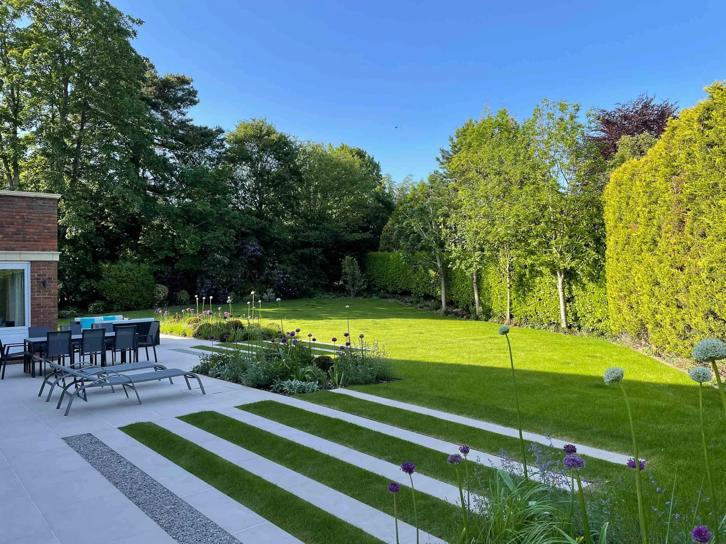 A backyard with a patio area featuring lounge chairs and a dining table with chairs. The lawn is well-maintained with a garden bed of flowers, and there are tall trees and a hedge lining the yard under a clear blue sky.