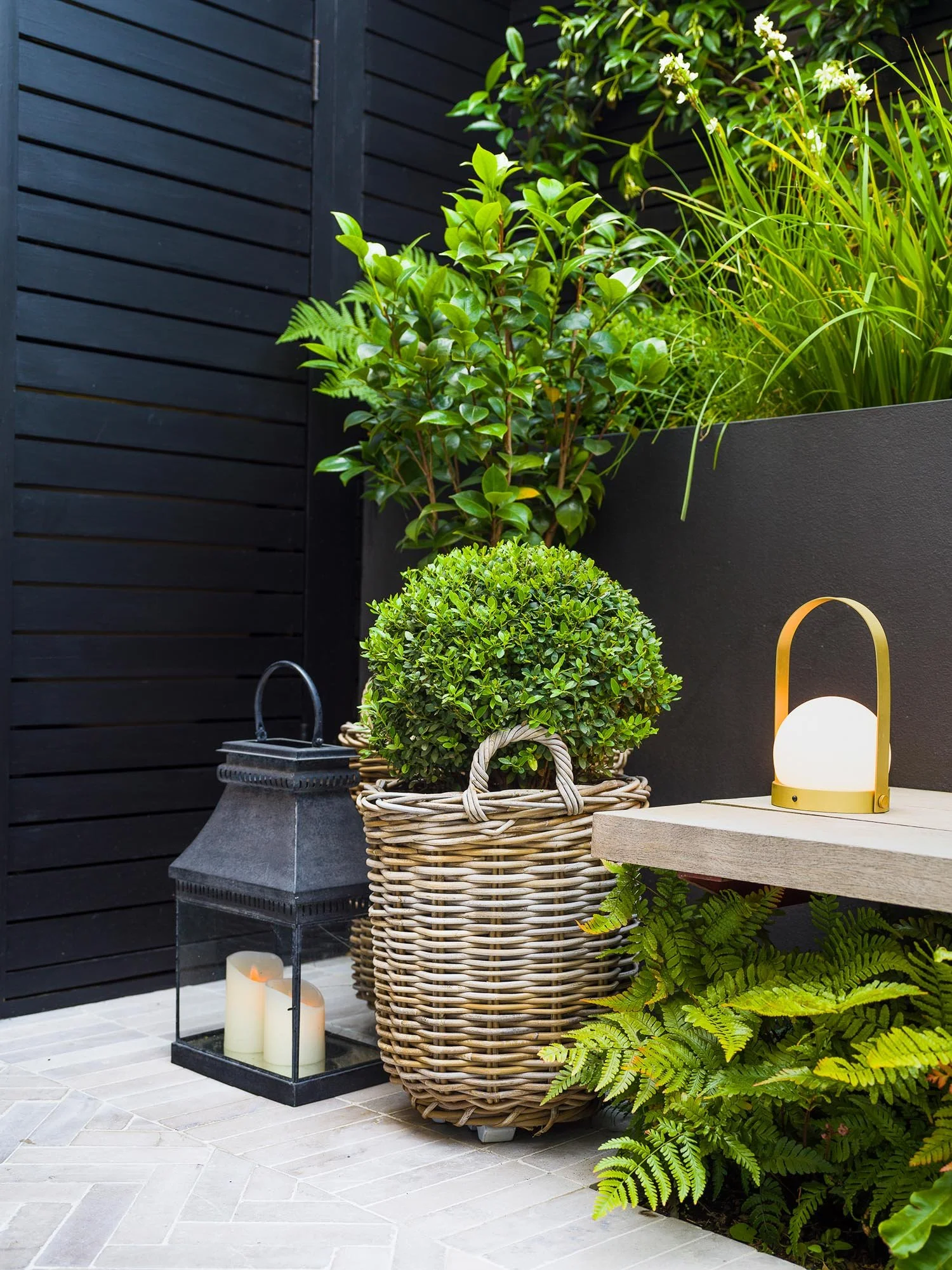 Outdoor corner with a woven basket planter containing a box ball next to a black lantern and a lit gold table lamp