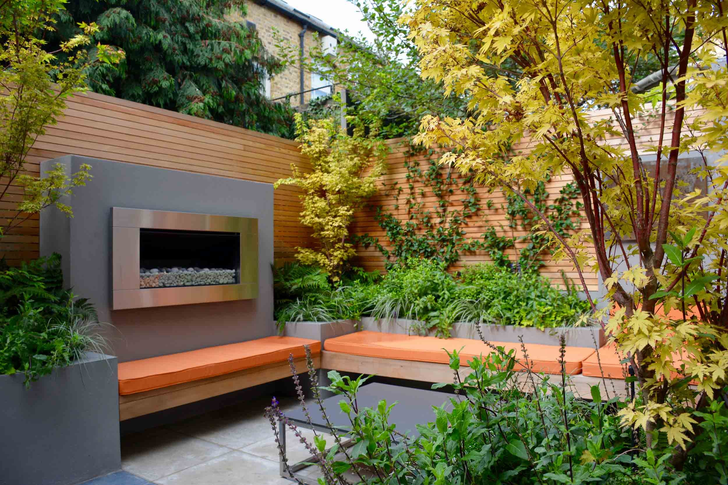 A cozy outdoor garden seating area with a modern fireplace, surrounded by lush green plants and trees, enclosed by a wooden privacy fence.