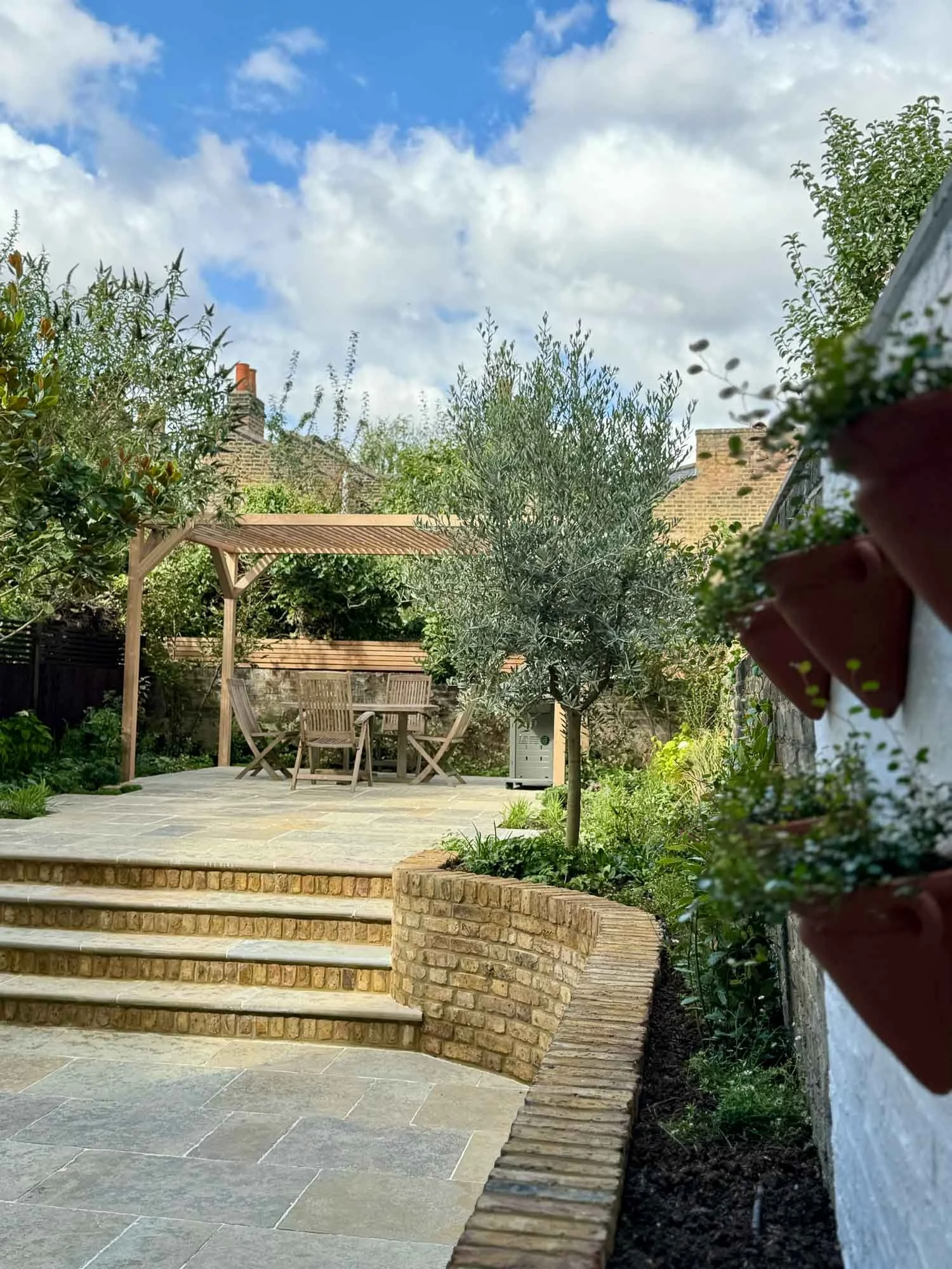 A well-maintained backyard garden scene with a patio, wooden table and chairs under a wooden pergola, small trees, potted plants on the side, and clear blue sky with scattered clouds.
