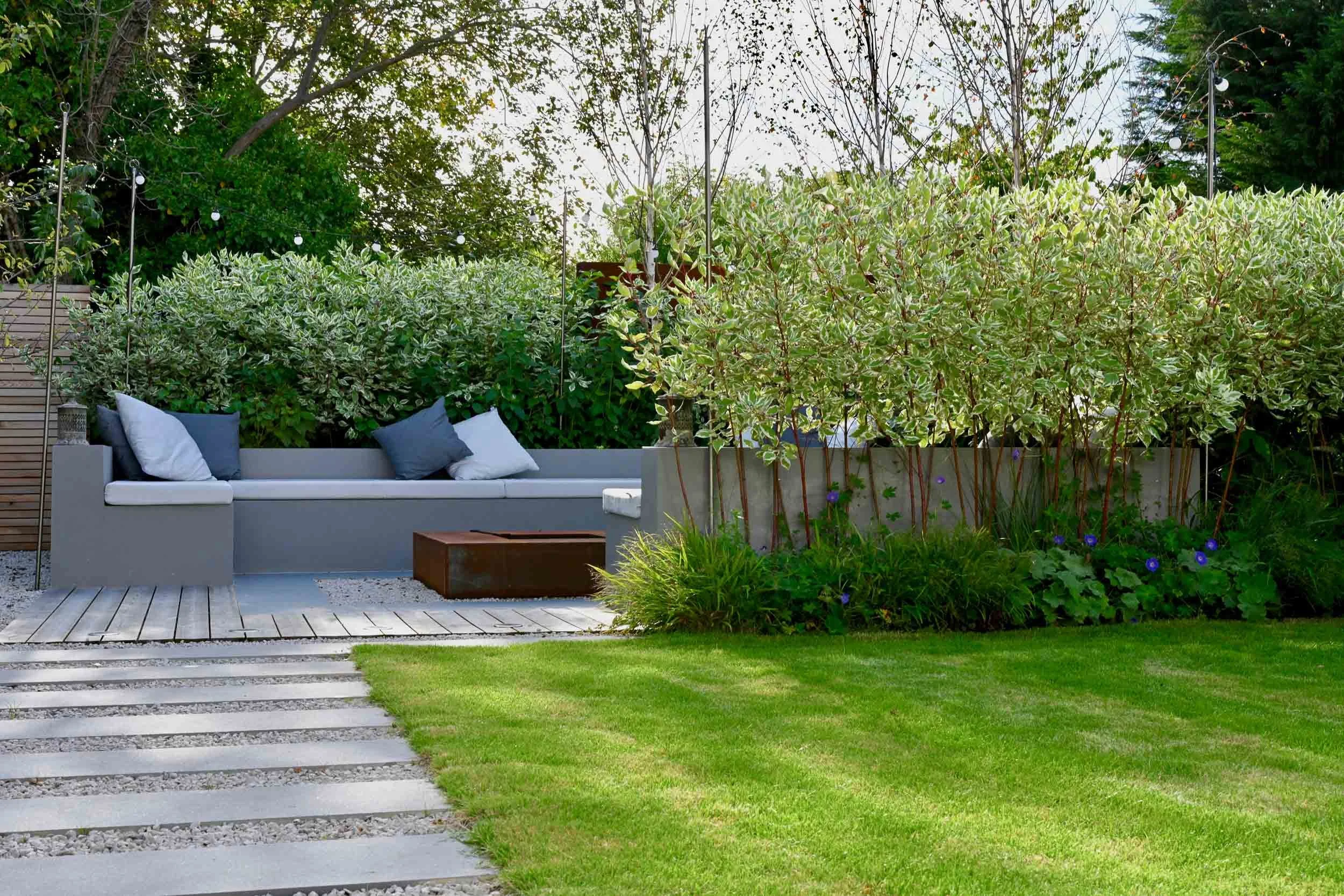 Modern backyard with a gray outdoor sofa, white and blue pillows, a rust-colored fire pit, green grass, and lush plants and trees, with string lights overhead.
