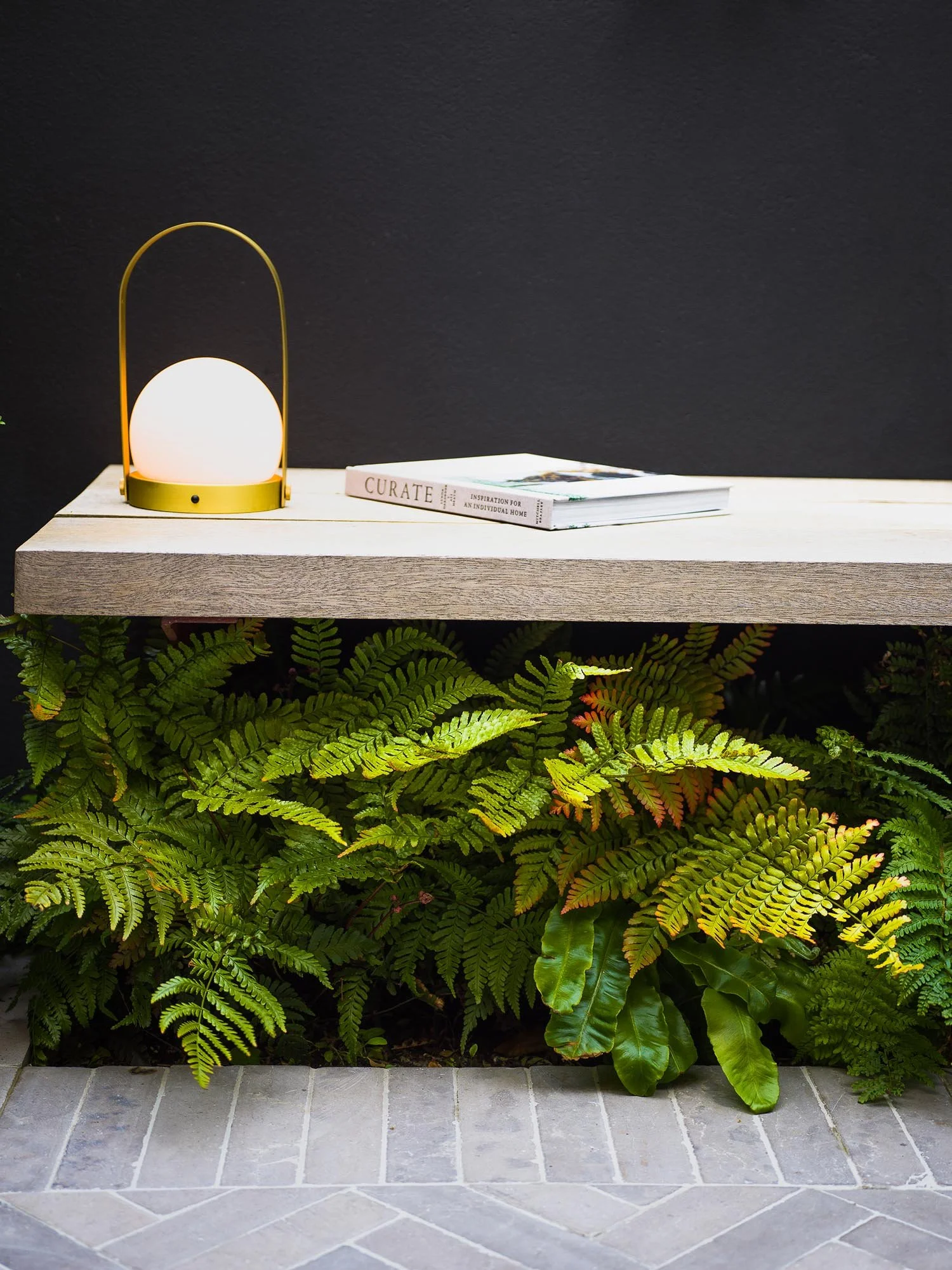 Close up of a floating oak bench with green ferns growing underneath and a brass lantern next to a white book