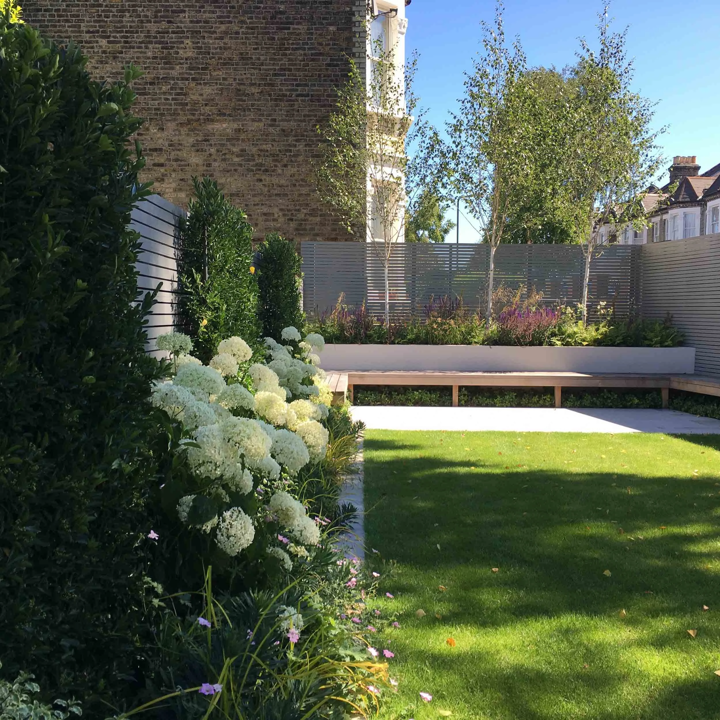A well-maintained backyard garden with green grass, white flowering bushes on the left, a raised flower bed with purple and green plants, and three small trees along a grey fence. The background includes a brick wall and blue sky.