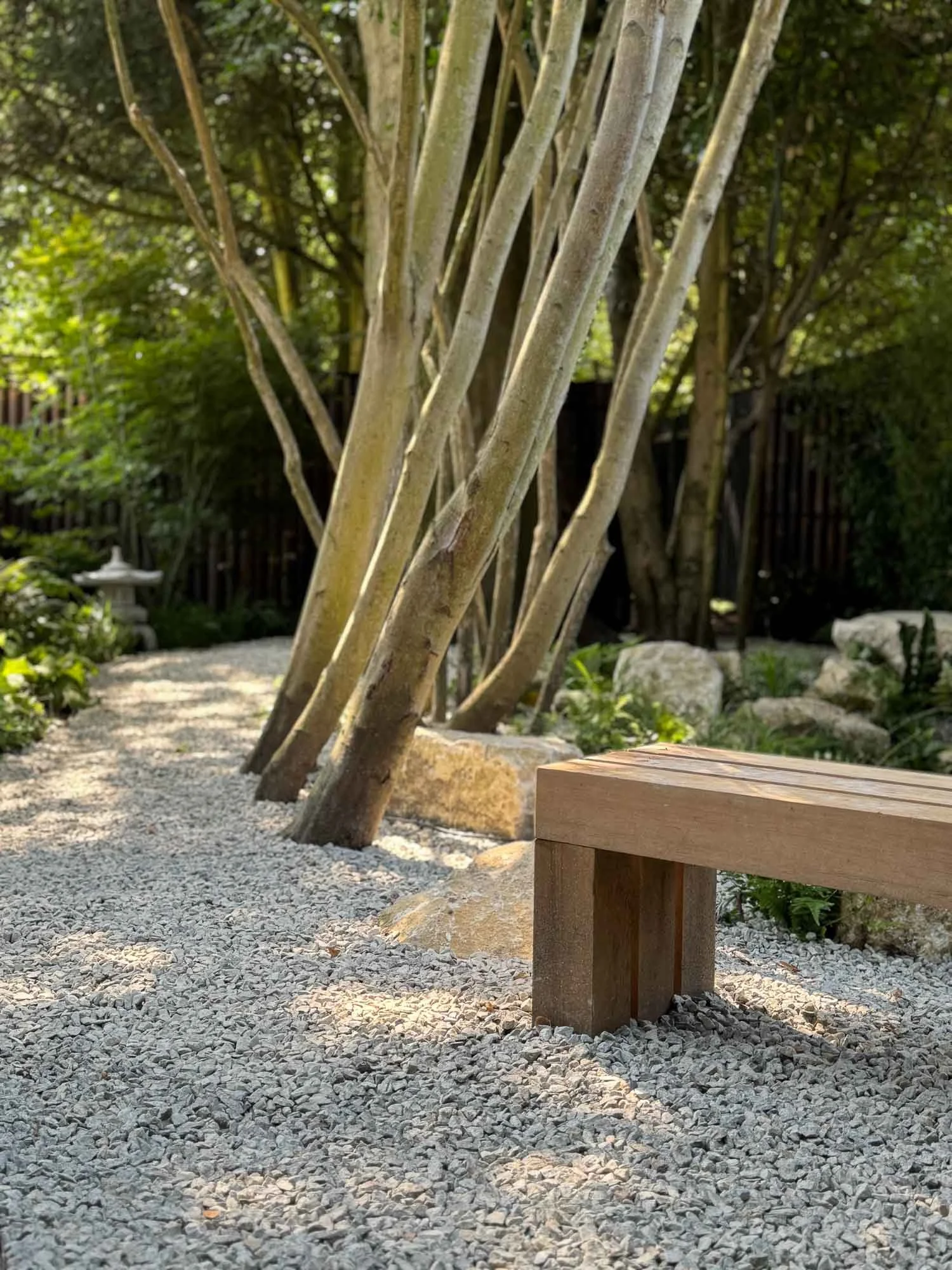 A wooden bench on gravel in a serene garden with trees, rocks, and greenery, along with a stone lantern in the background.