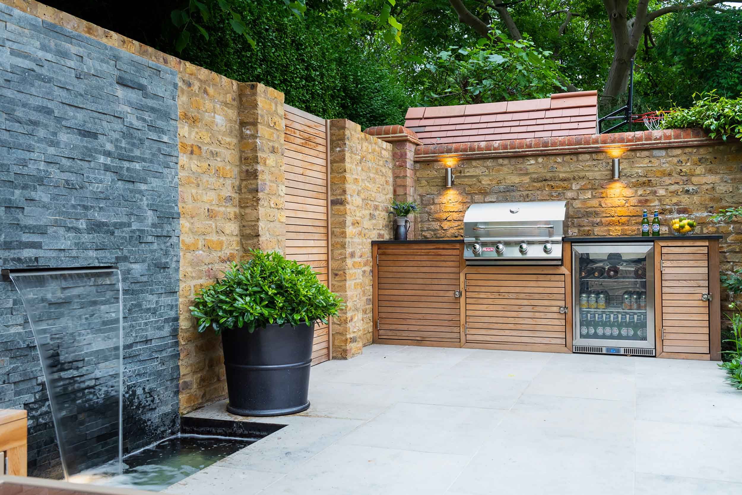 Garden patio showing a slate water feature and a bespoke outdoor kitchen with slatted timber doors and integrated grill