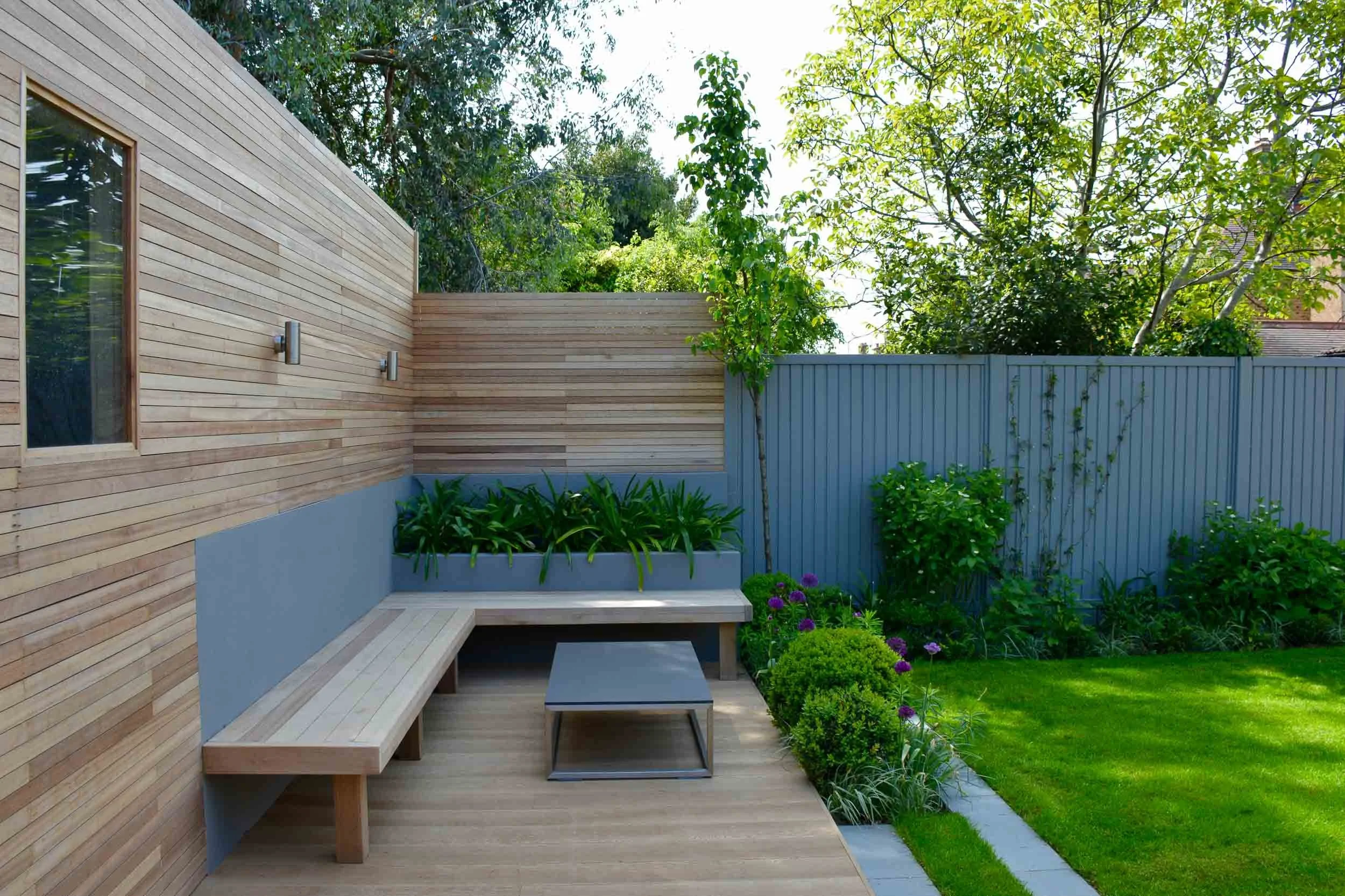 Modern backyard with wooden deck, built-in bench, garden plants, grass lawn, and fence, surrounded by trees.