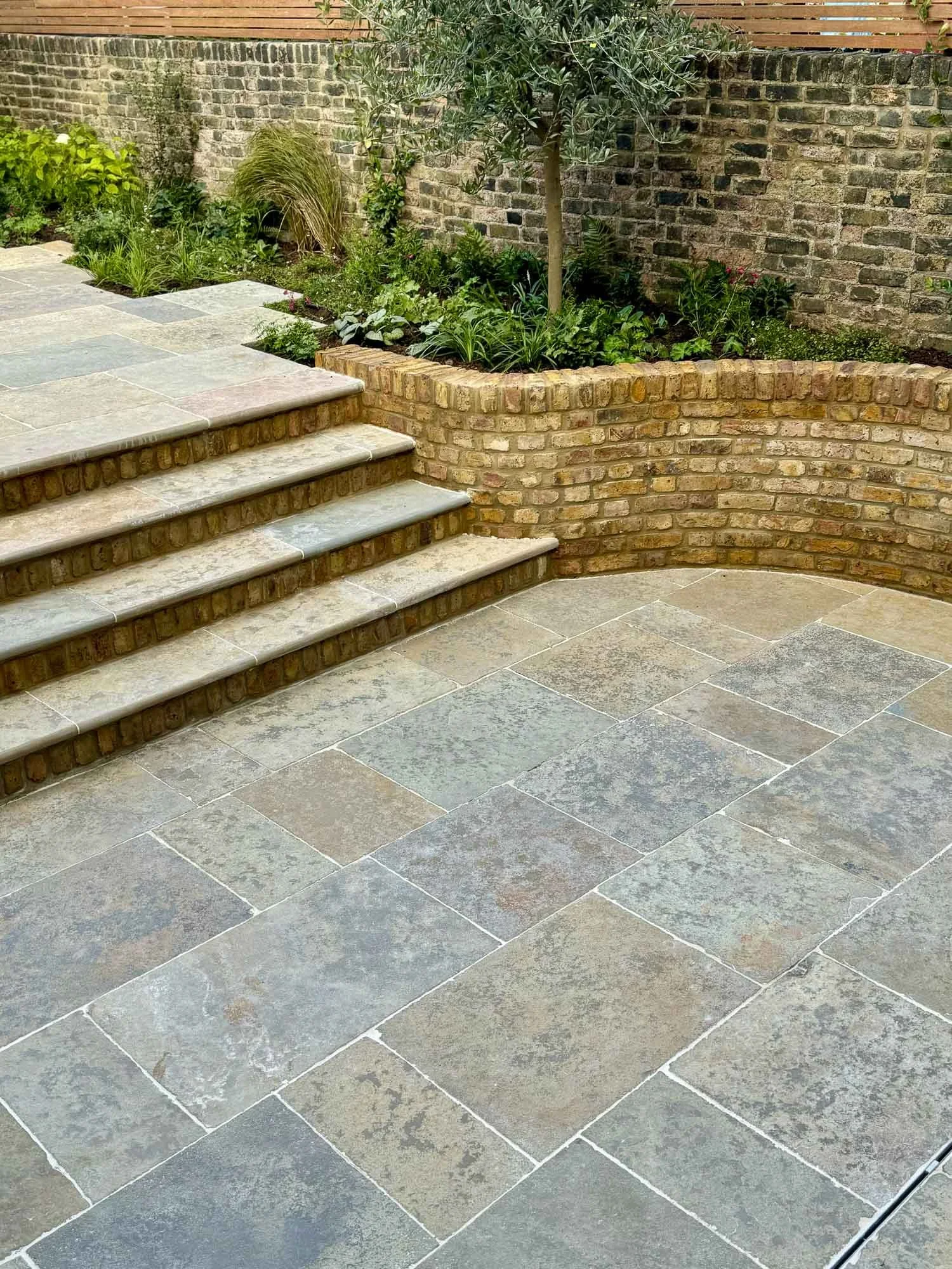 Stone patio with steps leading to a garden area, surrounded by a brick retaining wall and various plants, including a small tree.