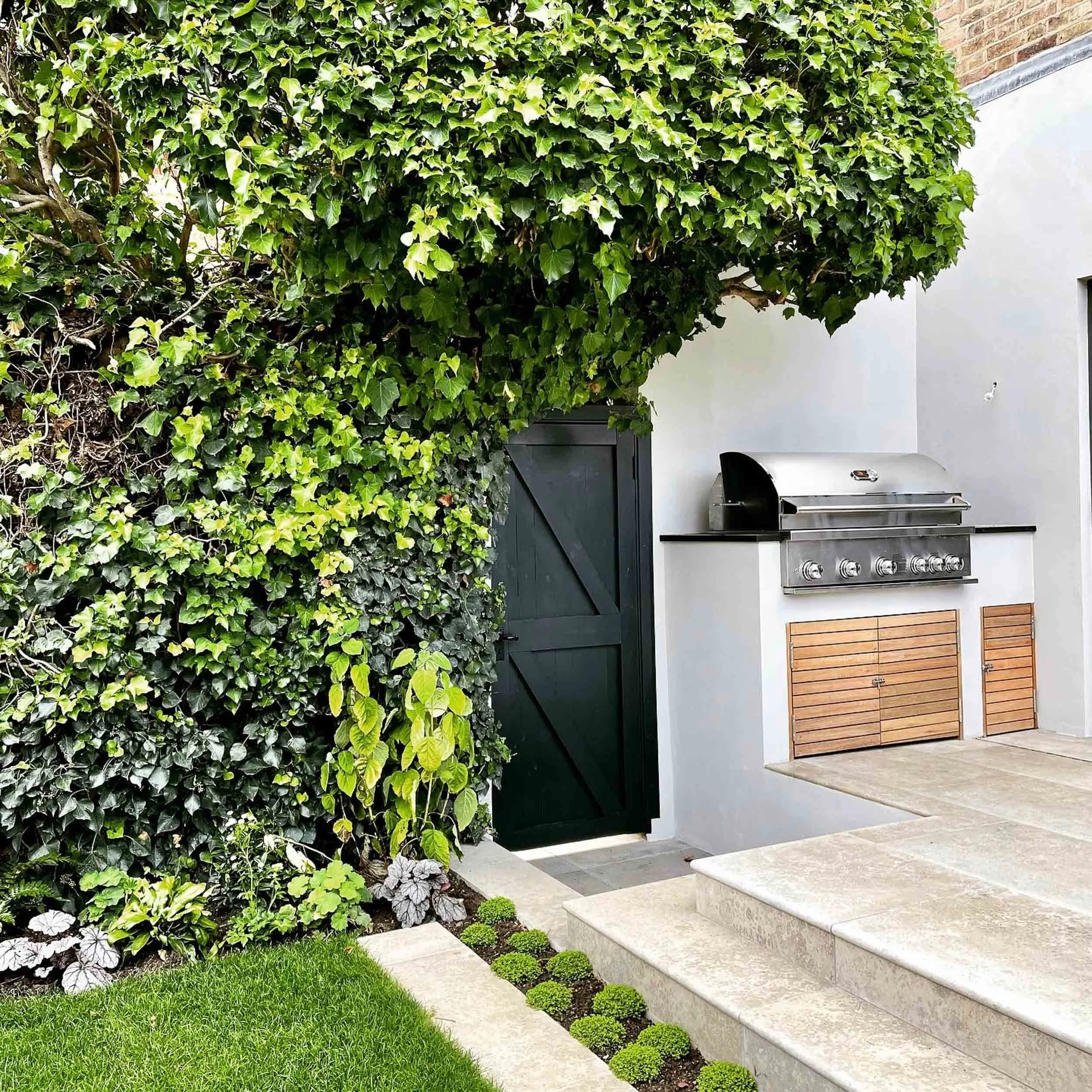 A backyard patio with a built-in stainless steel grill, white house wall, dark green wooden gate, lush green ivy-covered hedge, small potted plants, a step, and a striped wooden panel under the grill.