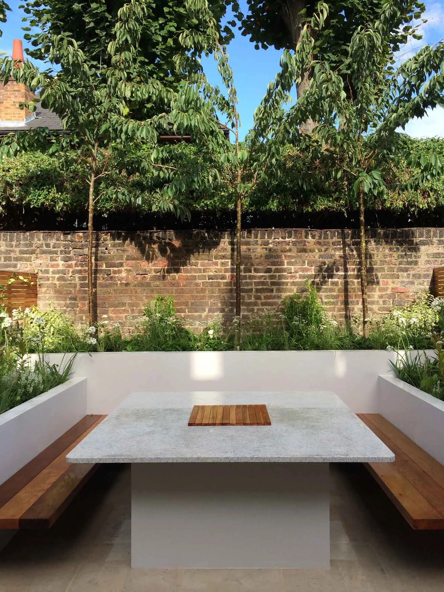 Outdoor seating area with a rectangular stone table, wooden benches, surrounding white planters with plants, a brick wall, trees, and a blue sky.