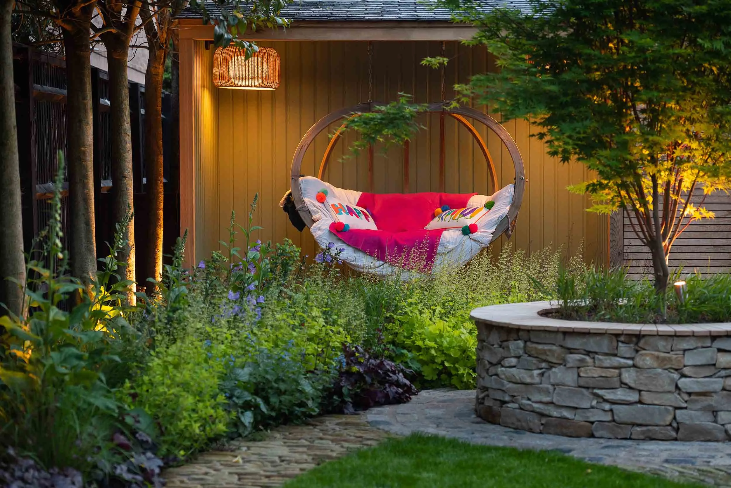 Illuminated garden at night with a glowing hanging chair and lit trees near a circular stone wall and planter