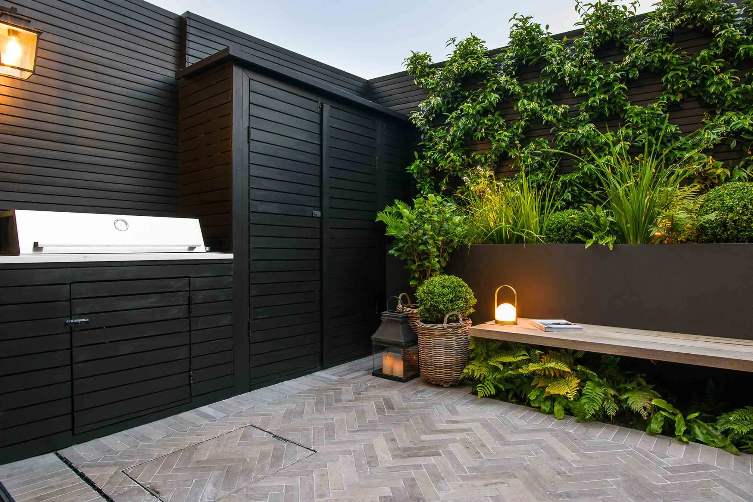 Outdoor kitchen and storage area with black timber cladding and a grey brick floor next to a lit wooden bench seat