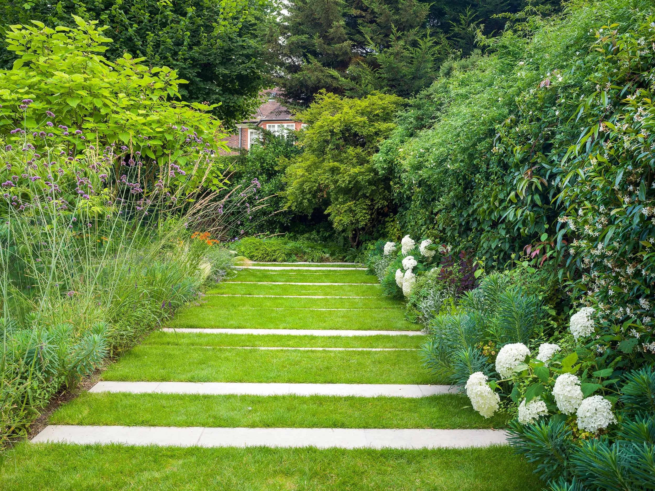 A lush green garden pathway with grass strips and stone tiles, bordered by various flowering plants and dense trees.