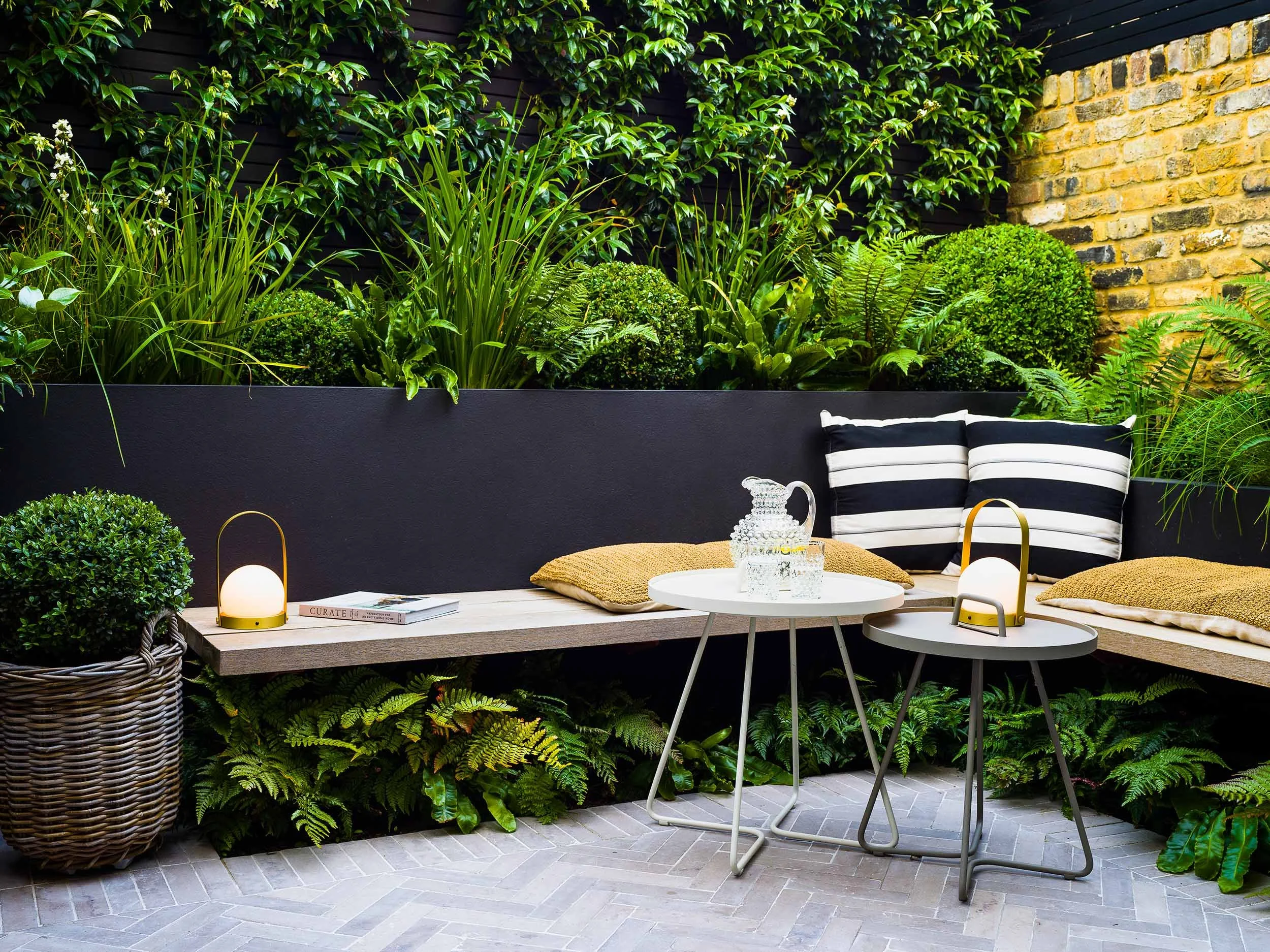 Outdoor patio seating area with a built-in black bench, yellow cushions, black-and-white striped pillows, small white round tables, decorative bottles, book, and small lamps, surrounded by lush green plants and foliage, with brick and black walls.