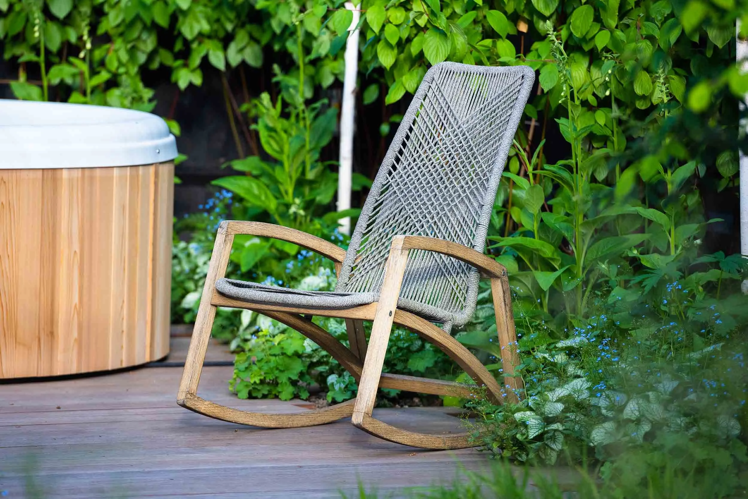 Grey woven rocking chair on a timber deck beside a wooden hot tub and lush green garden foliage with blue flowers