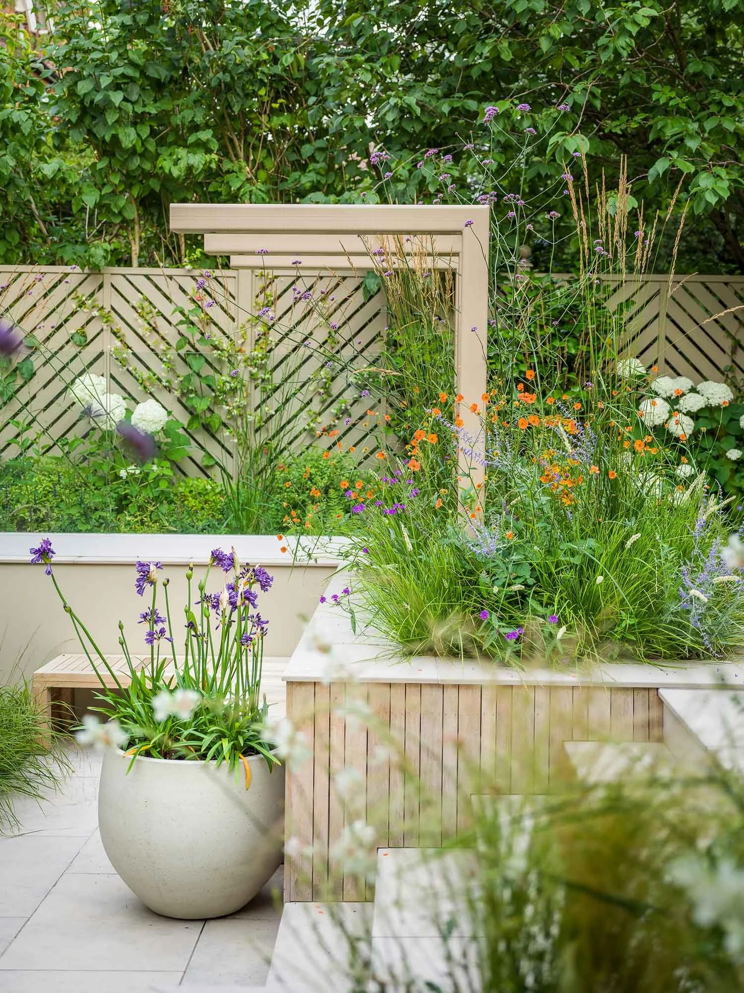 Garden detail showing a beige pergola structure over a raised planter bed and a large pot of blue Agapanthus
