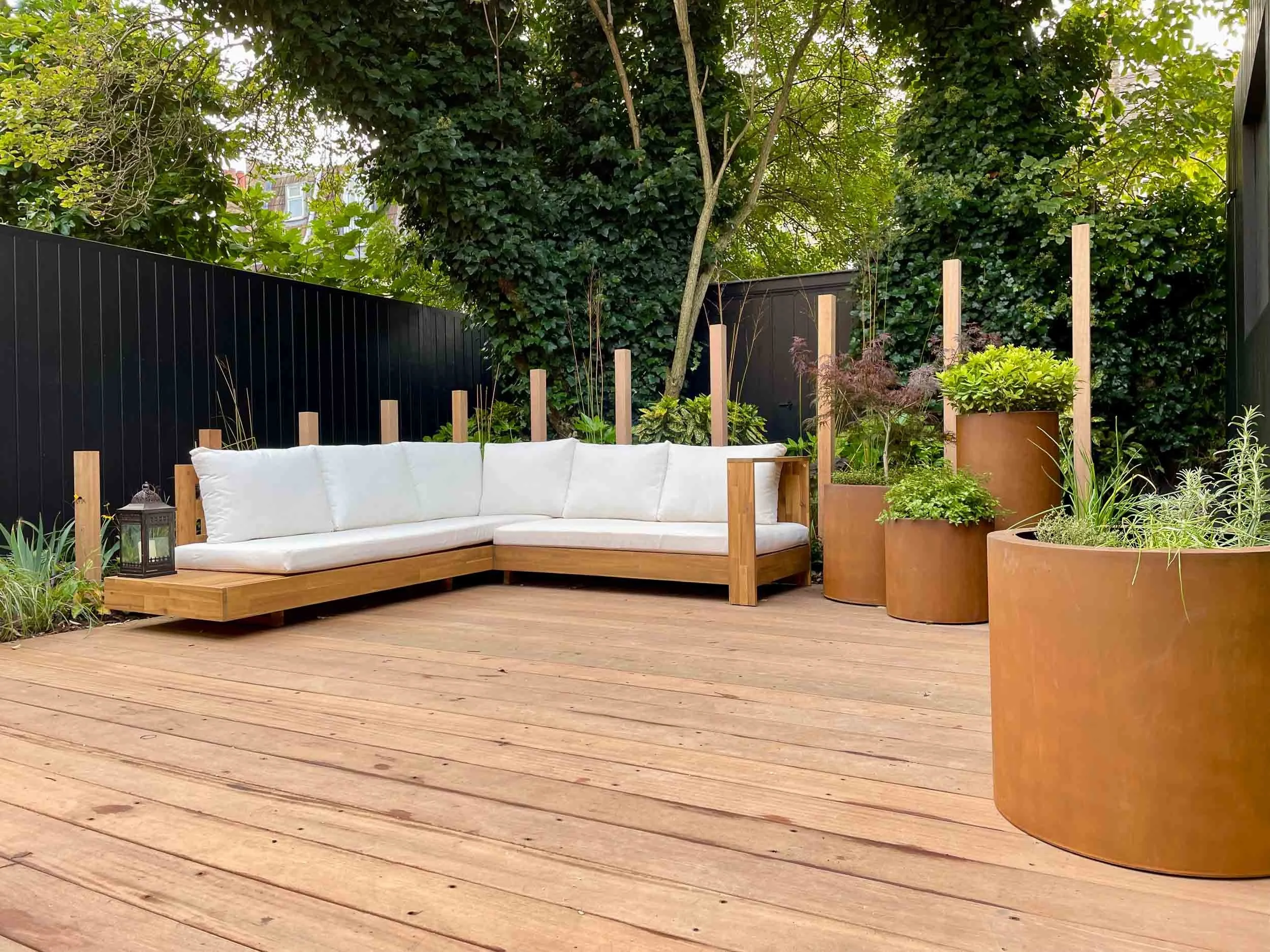Outdoor patio with wooden deck, white cushioned corner sofa, large rust-colored plant containers with various green plants, black fencing, and lush green trees and shrubs in the background.