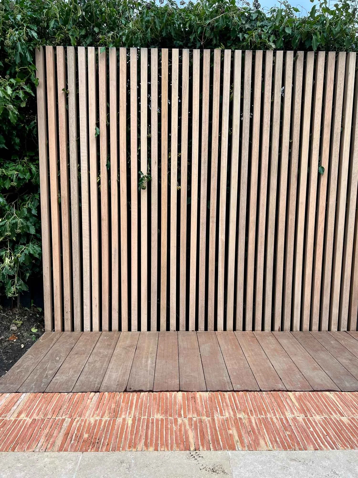 Wooden vertical slat privacy fence with wooden deck below and brick paving in front, greenery in the background.