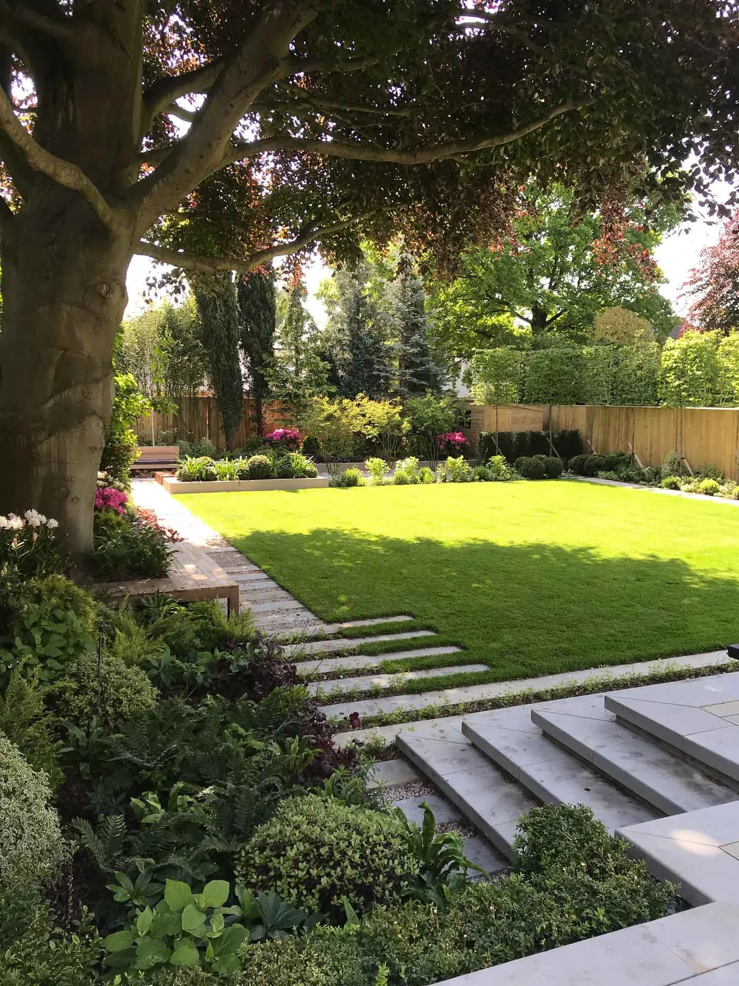 A lush backyard garden with a large tree, a green lawn, paved stone walkways, flower beds, and a wooden fence.